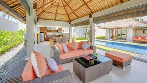 A covered outdoor seating area with wicker furniture, a pool, and a modern kitchen in the background. - Villa Jerami & Spa (Seminyak)