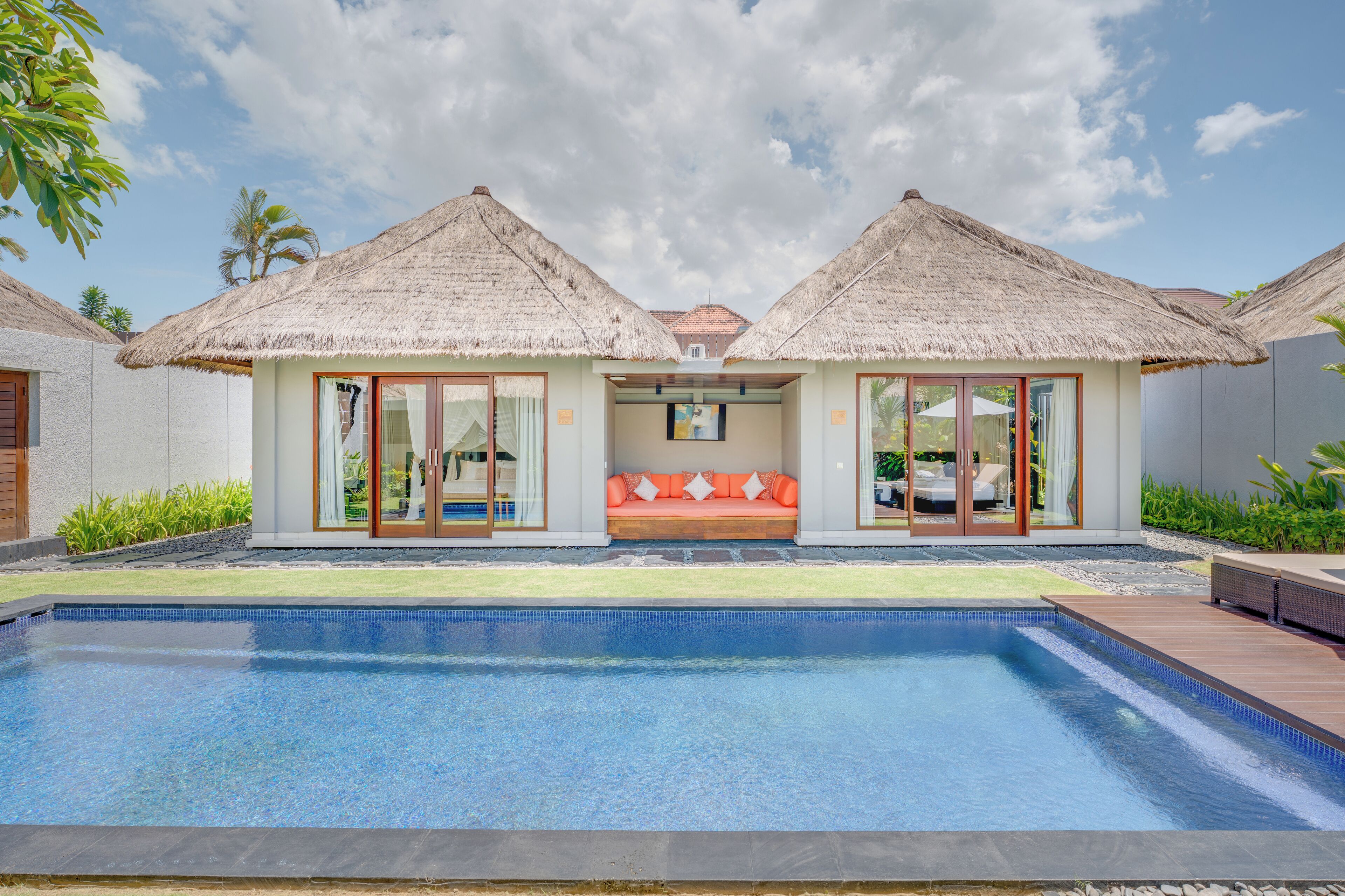 A resort with a swimming pool, two thatched-roof structures, and a seating area with cushions.