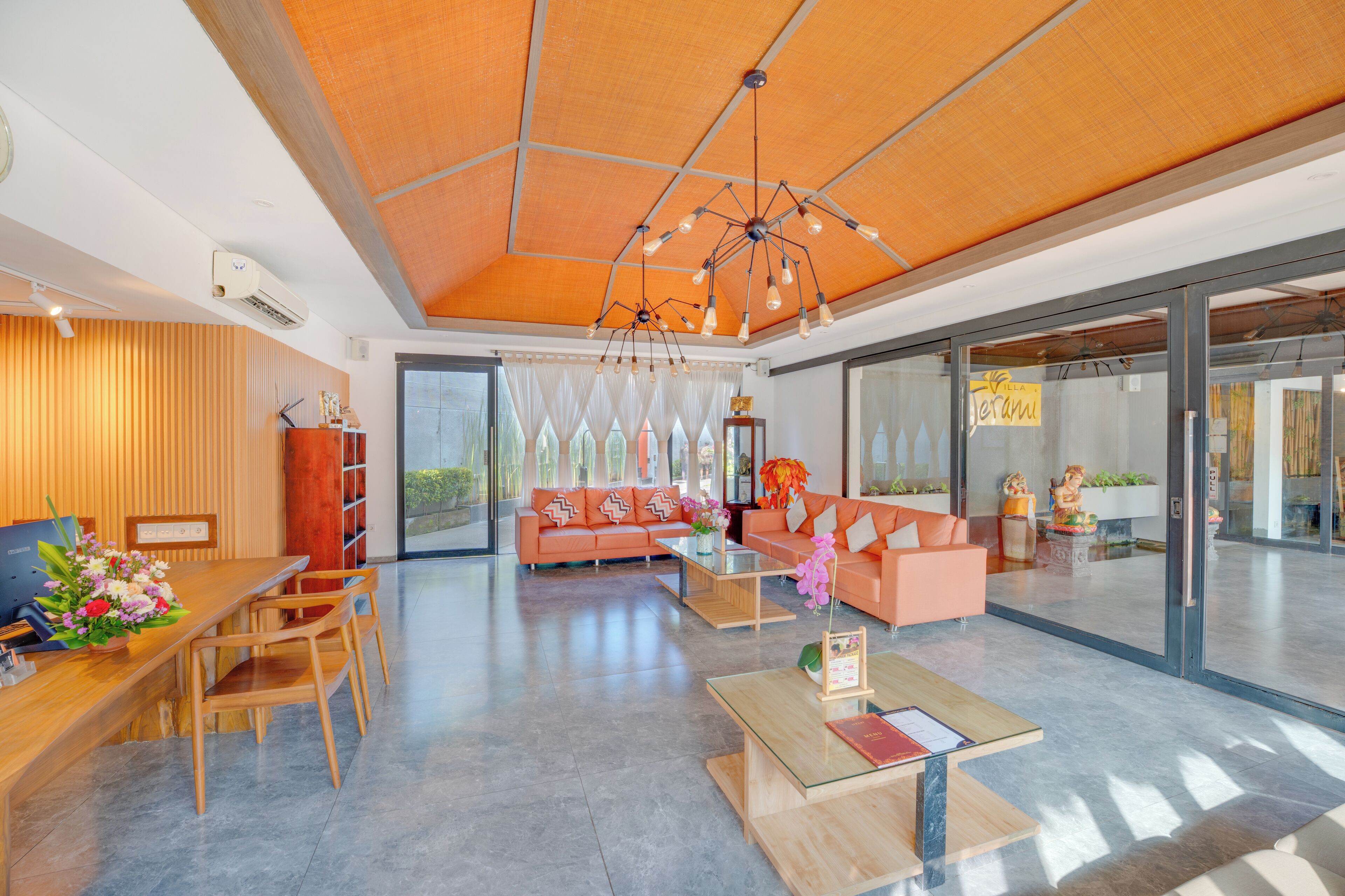 A spacious lobby with a wooden reception desk, a sofa, and a coffee table. There is a large window with sheer curtains and a decorative plant.