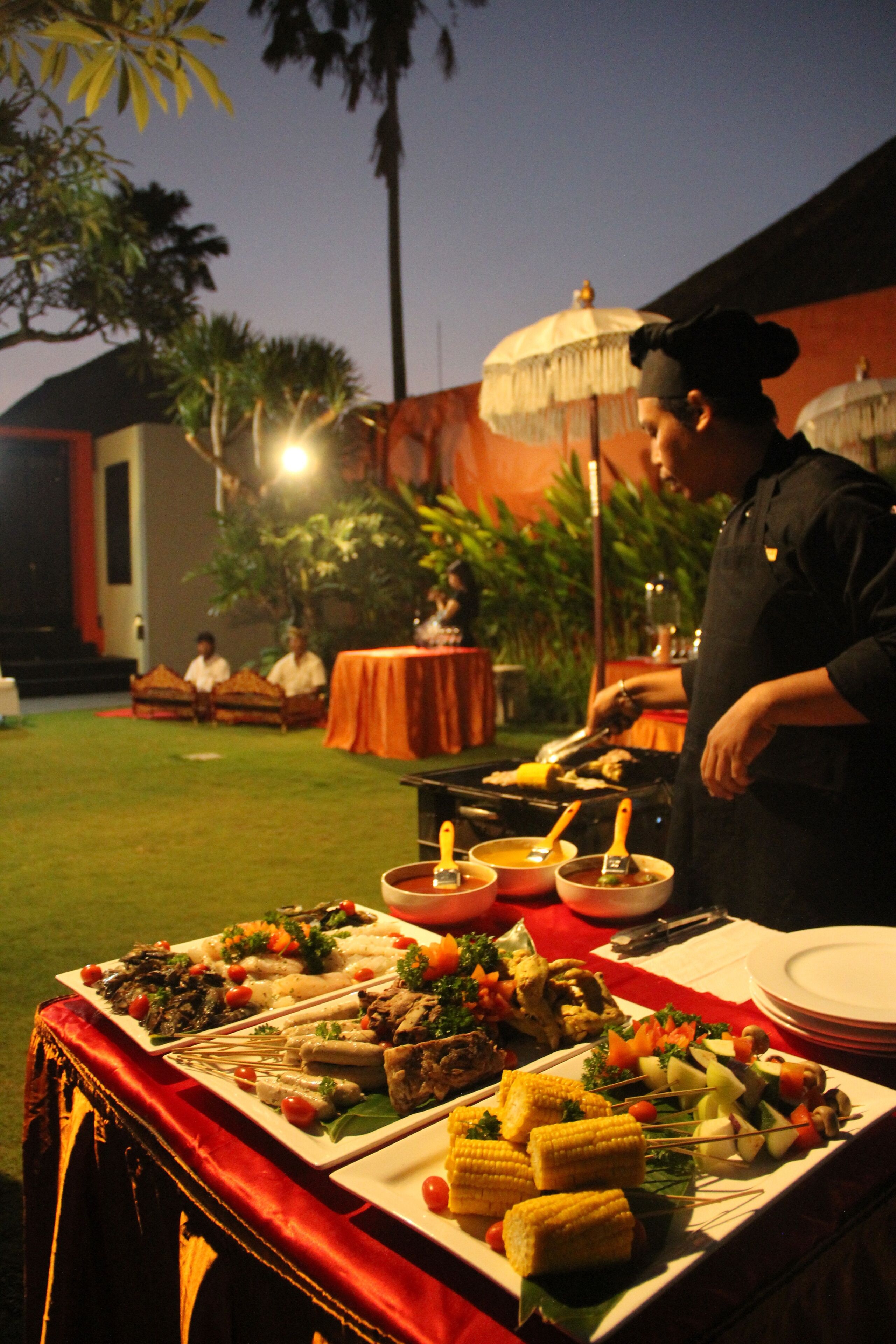 A buffet setup with various dishes, including corn on the cob and skewers, in an outdoor setting with palm trees and a building in the background.