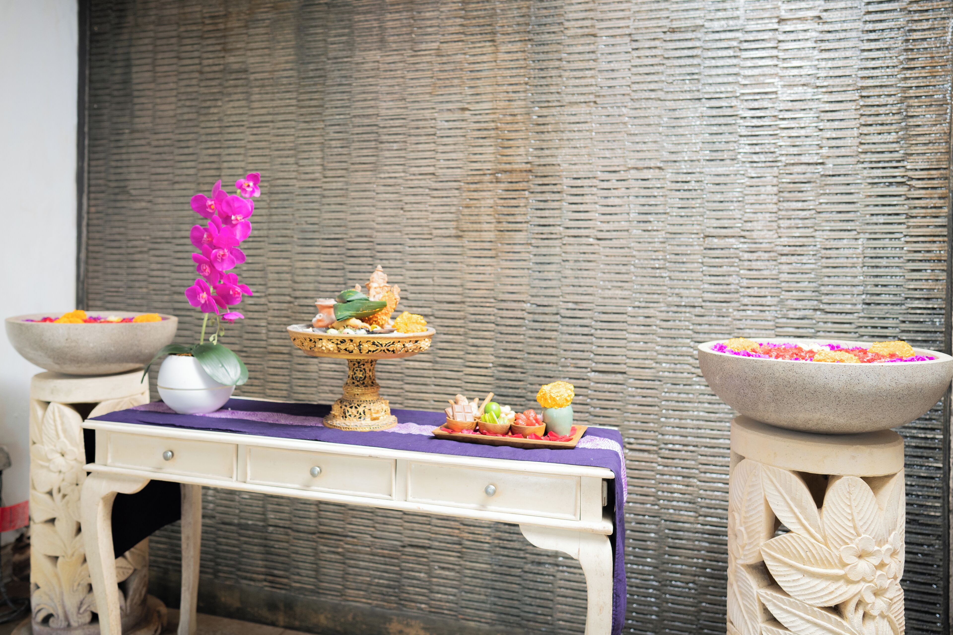 A table with a purple cloth, a white vase with a purple orchid, and two large bowls with colorful flowers.