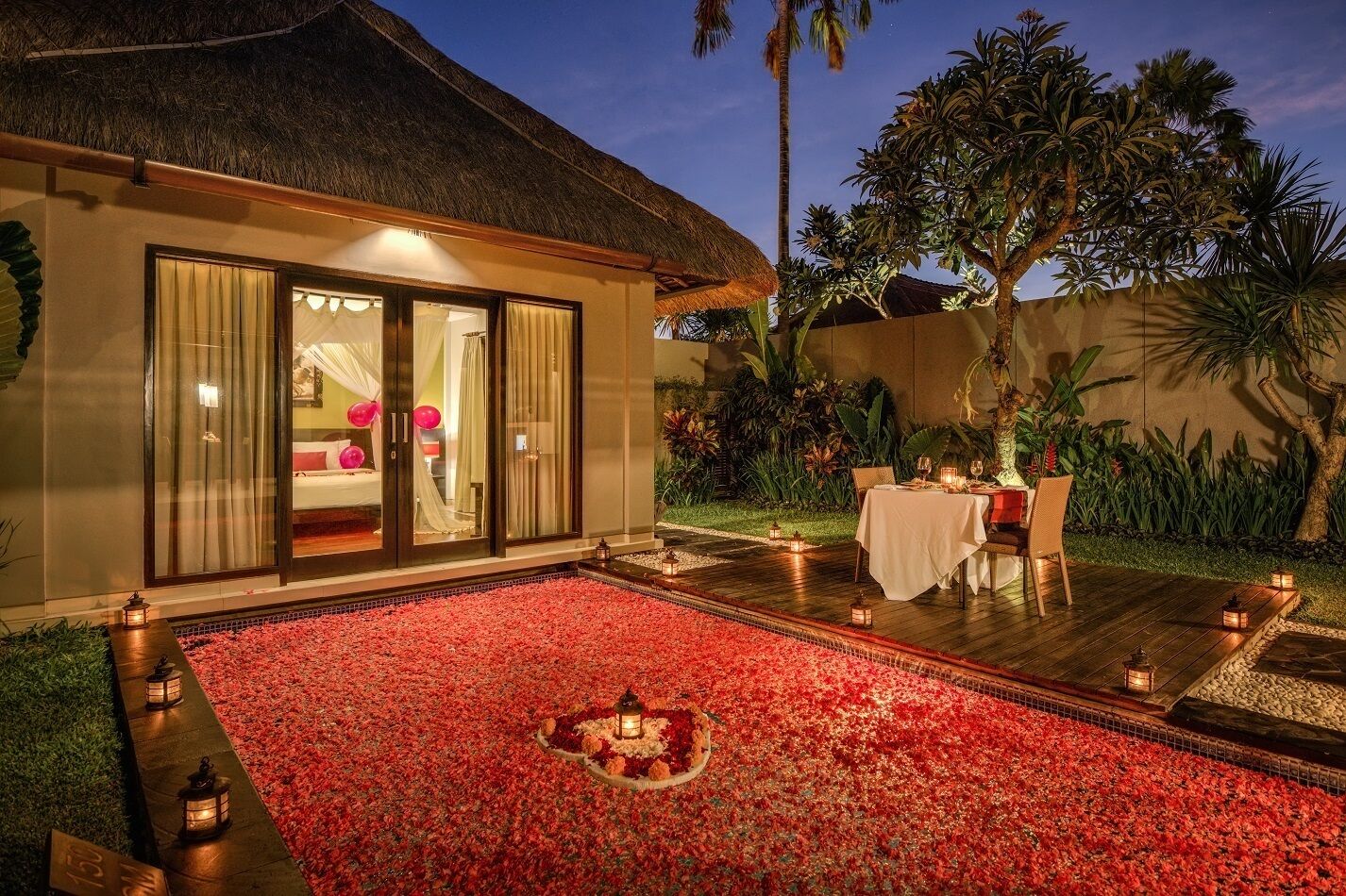 A private villa with a glass-enclosed bedroom, a dining area with a table set for two, and a red flower carpet leading to the entrance.
