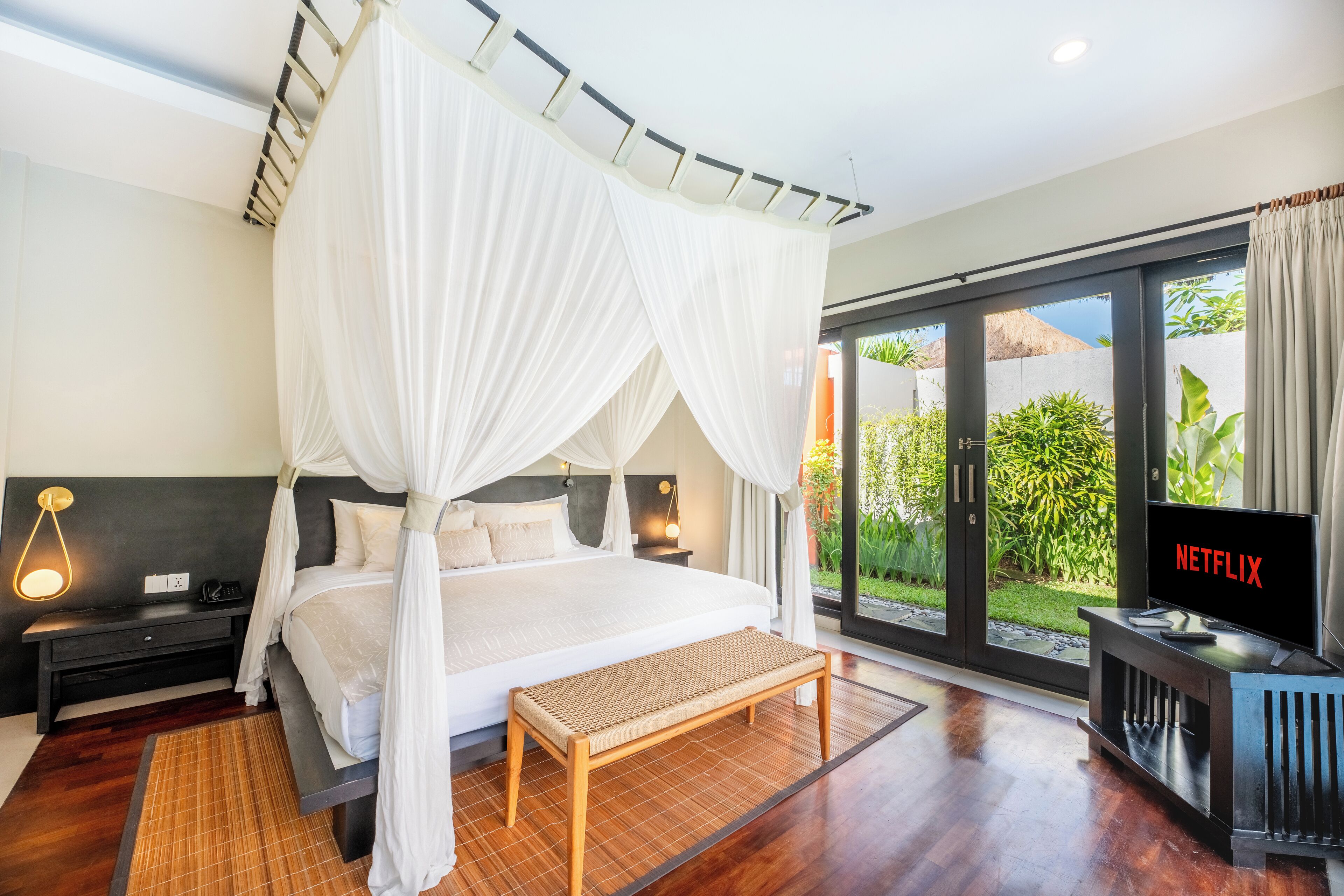 A bedroom with a canopy bed, a bench, a TV, and a view of greenery outside.