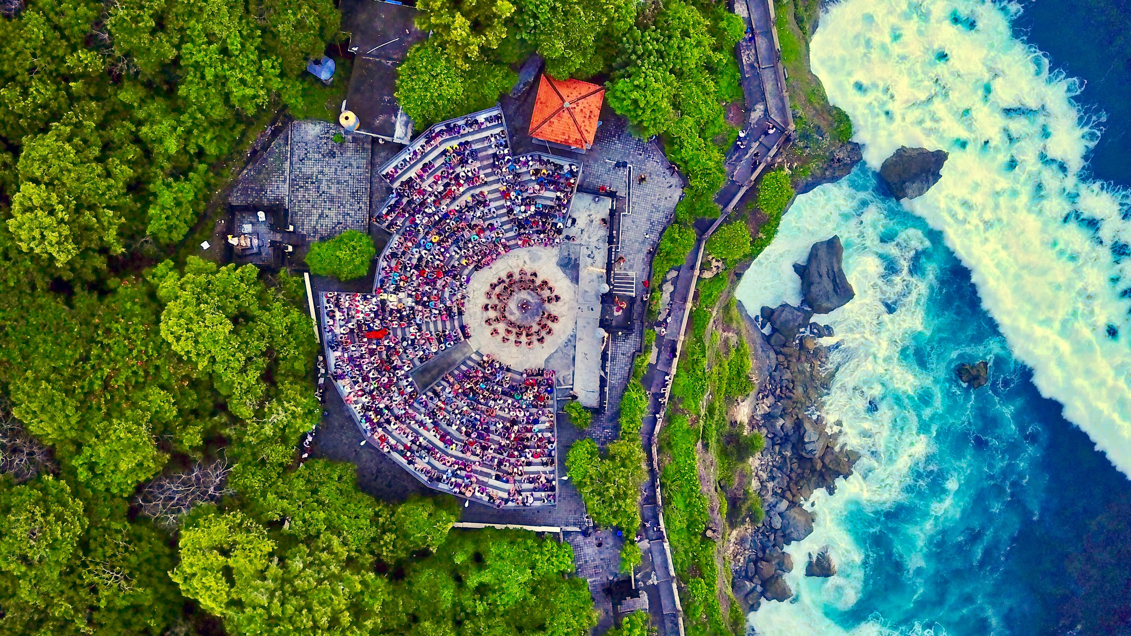 An aerial view of a circular amphitheater with tiered seating, surrounded by lush greenery and a rocky coastline.