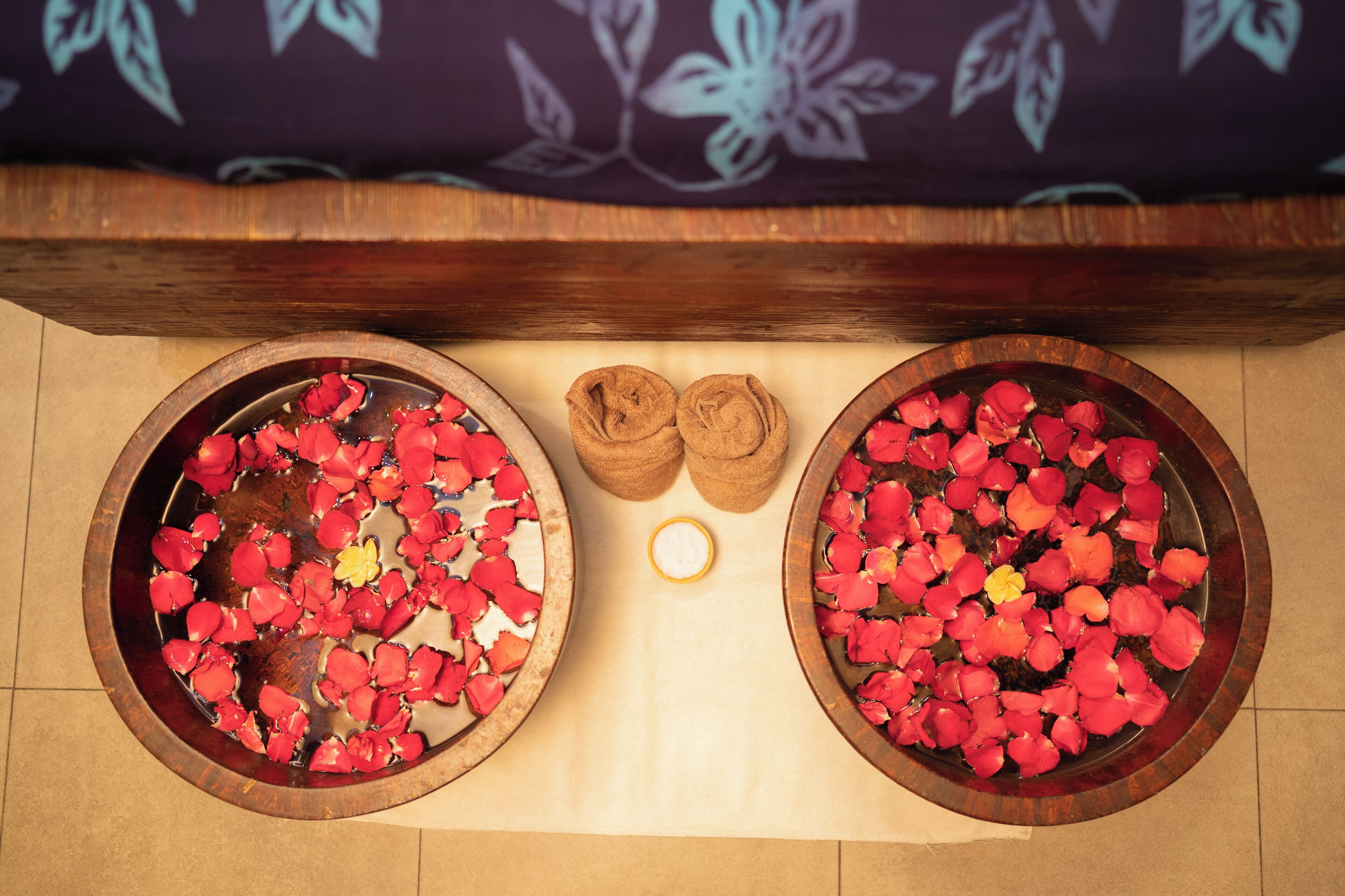 Two wooden bowls filled with rose petals and a candle on a tiled floor.