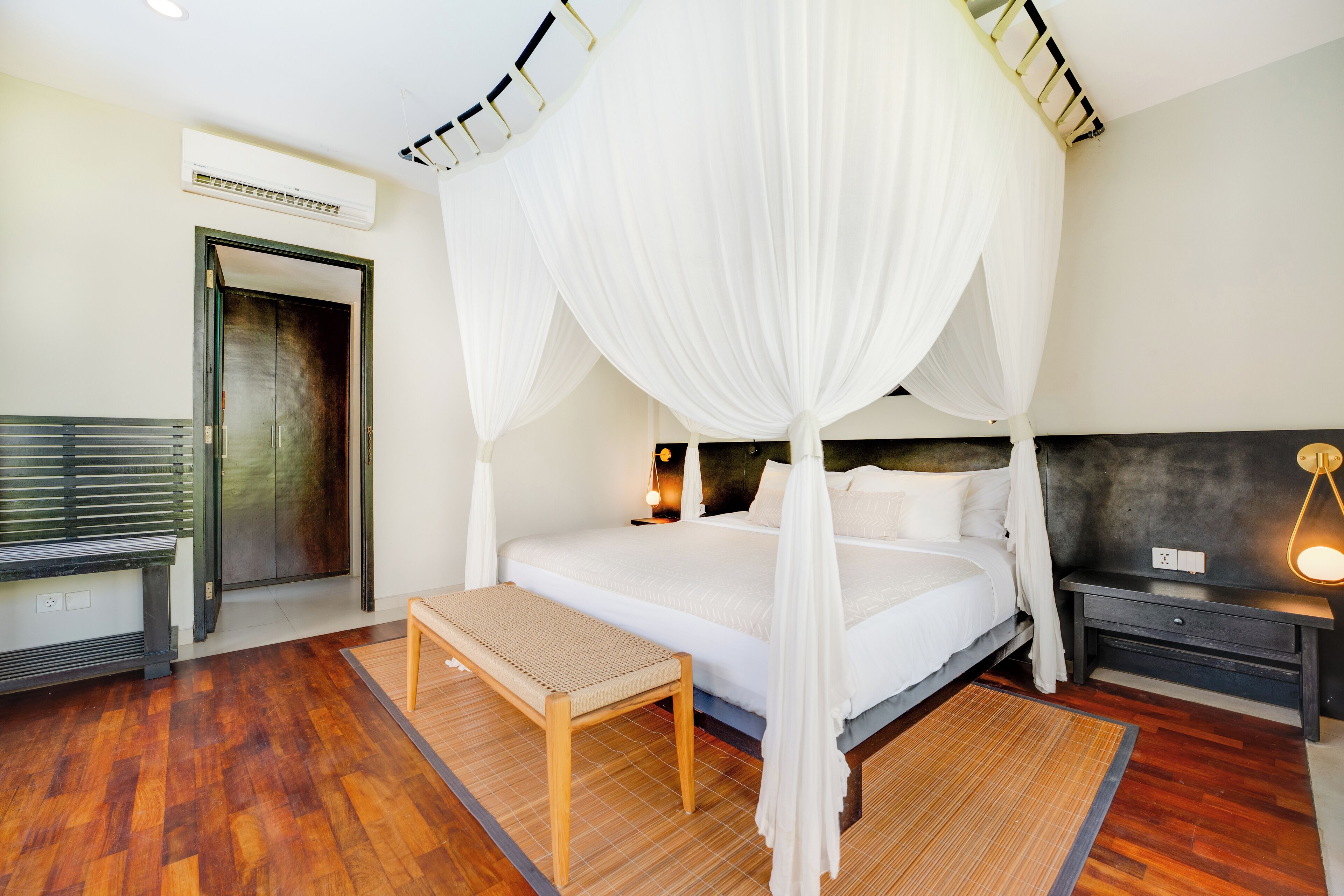 A bedroom with a four-poster bed, a bench, a bedside table, and a wall-mounted air conditioner.