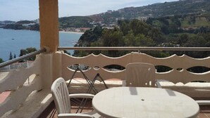 A balcony with a table and chairs, overlooking a coastal landscape with hills and a body of water.