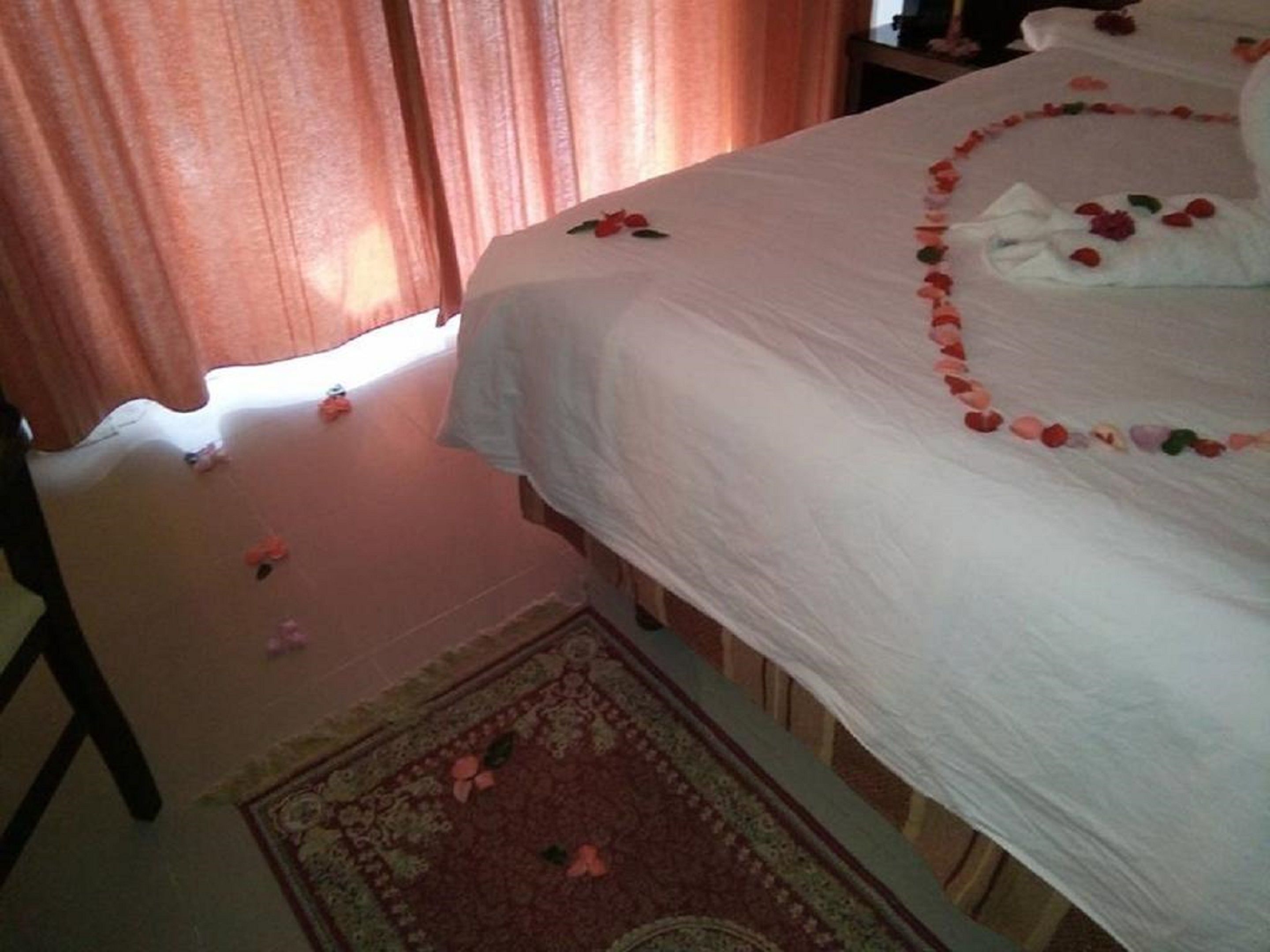 A neatly made bed with rose petals arranged in a heart shape, a white bathrobe, and a small rug with a floral pattern.