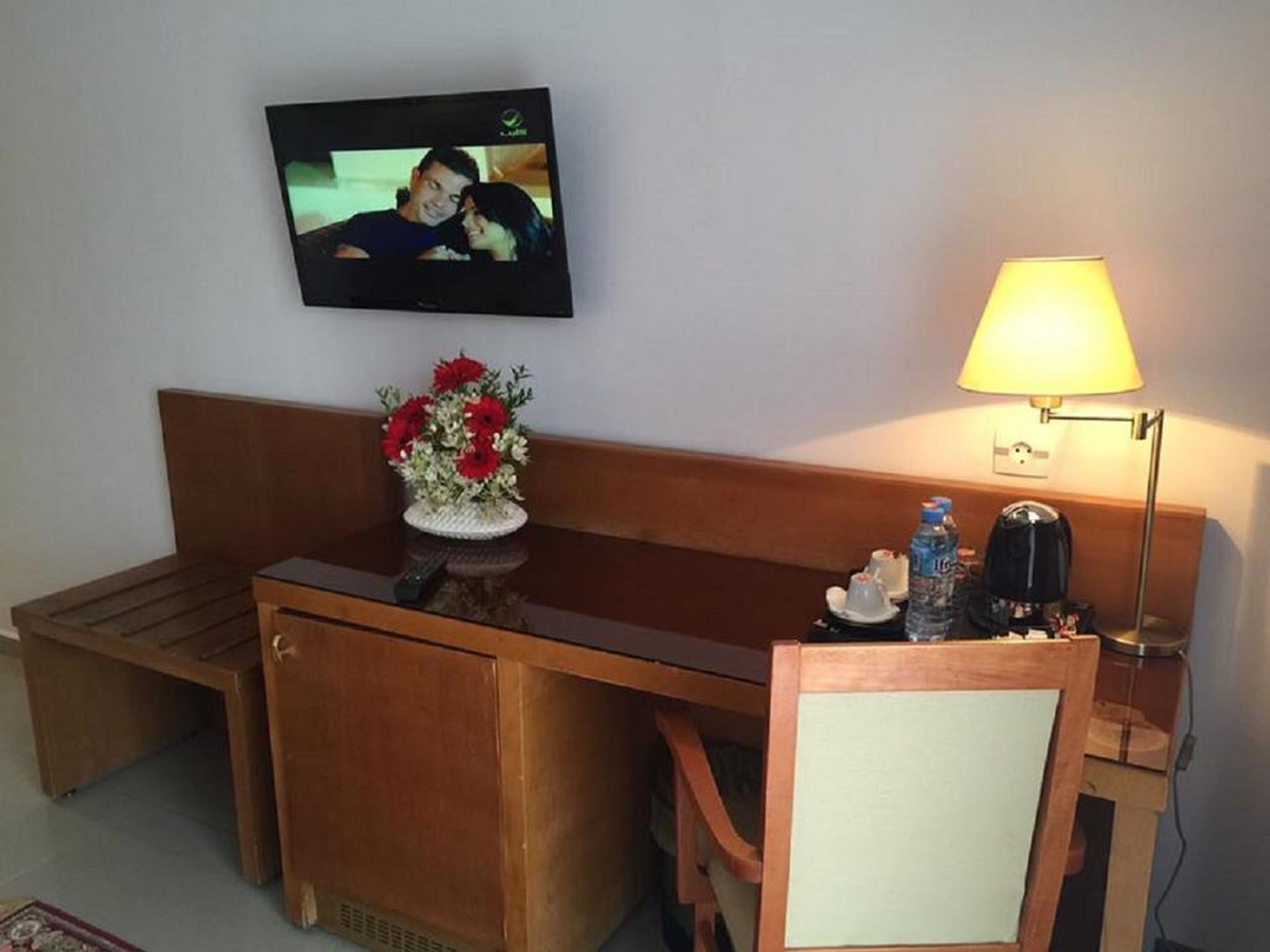 Habitación de hotel con un banco de madera, una mesita con un florero de flores rojas, un televisor montado en la pared y una lámpara de noche.