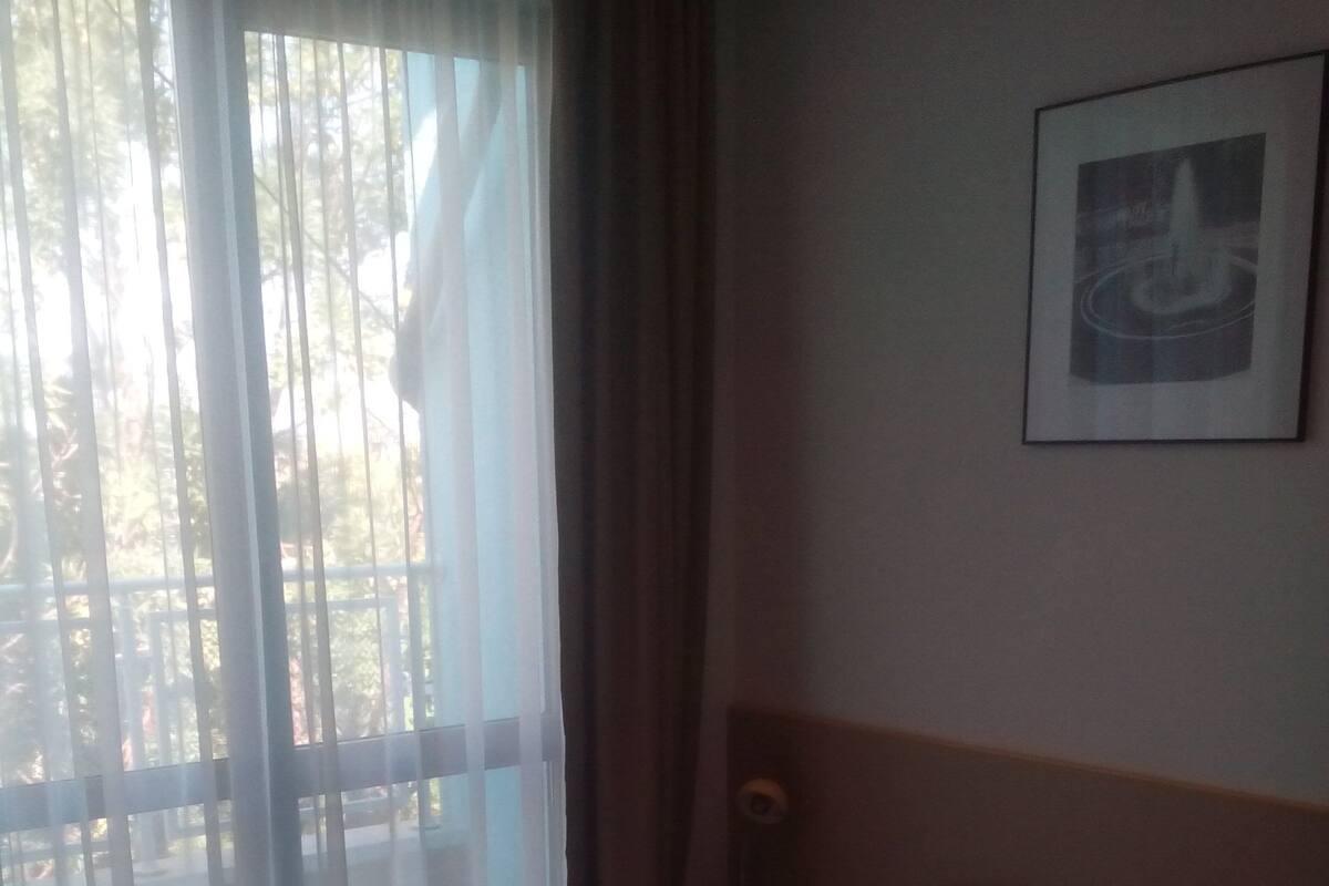 Une chambre d’hôtel avec un lit, une fenêtre avec des rideaux légers et un tableau encadré au mur.