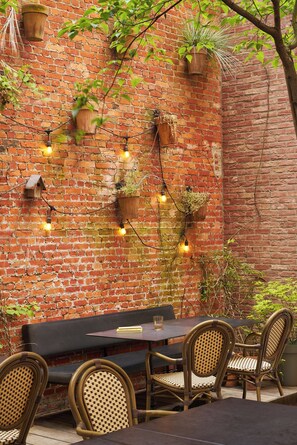 Un espace extérieur aménagé pour les repas, avec un mur en briques, des jardinières suspendues, des guirlandes lumineuses et des chaises en osier.