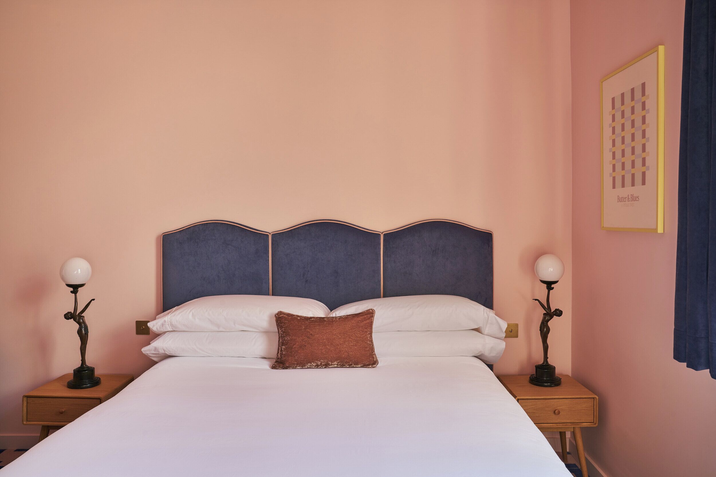 A neatly made bed with white and brown pillows, flanked by two bedside tables with lamps, in a room with a peach-colored wall and a framed artwork.