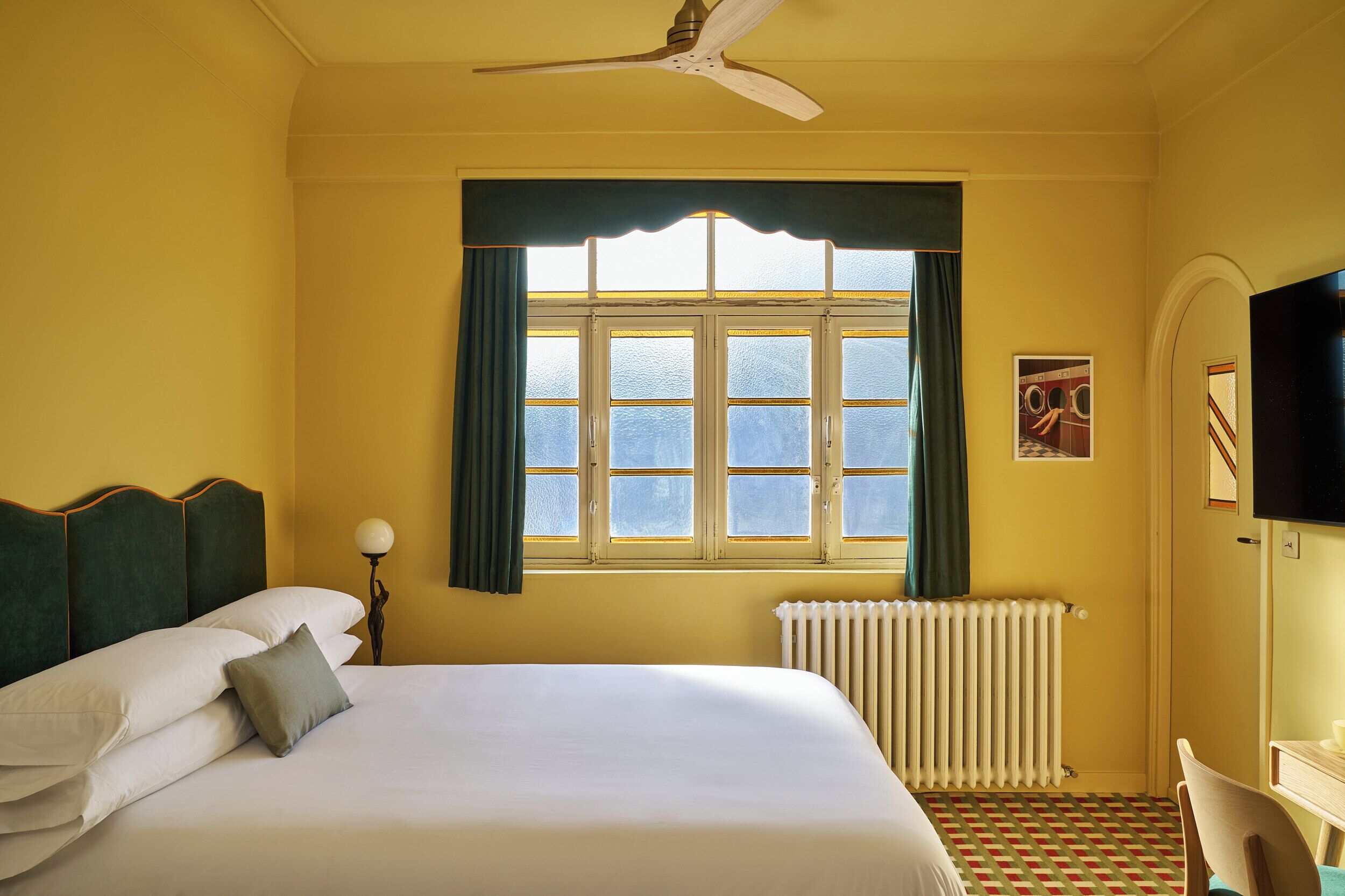 Camera d'albergo con un letto, un termosifone, una finestra con tende e un ventilatore a soffitto, oltre a una TV fissata al muro.