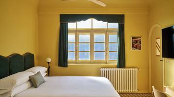 Camera d'albergo con un letto, un termosifone, una finestra con tende e un ventilatore a soffitto, oltre a una TV fissata al muro.