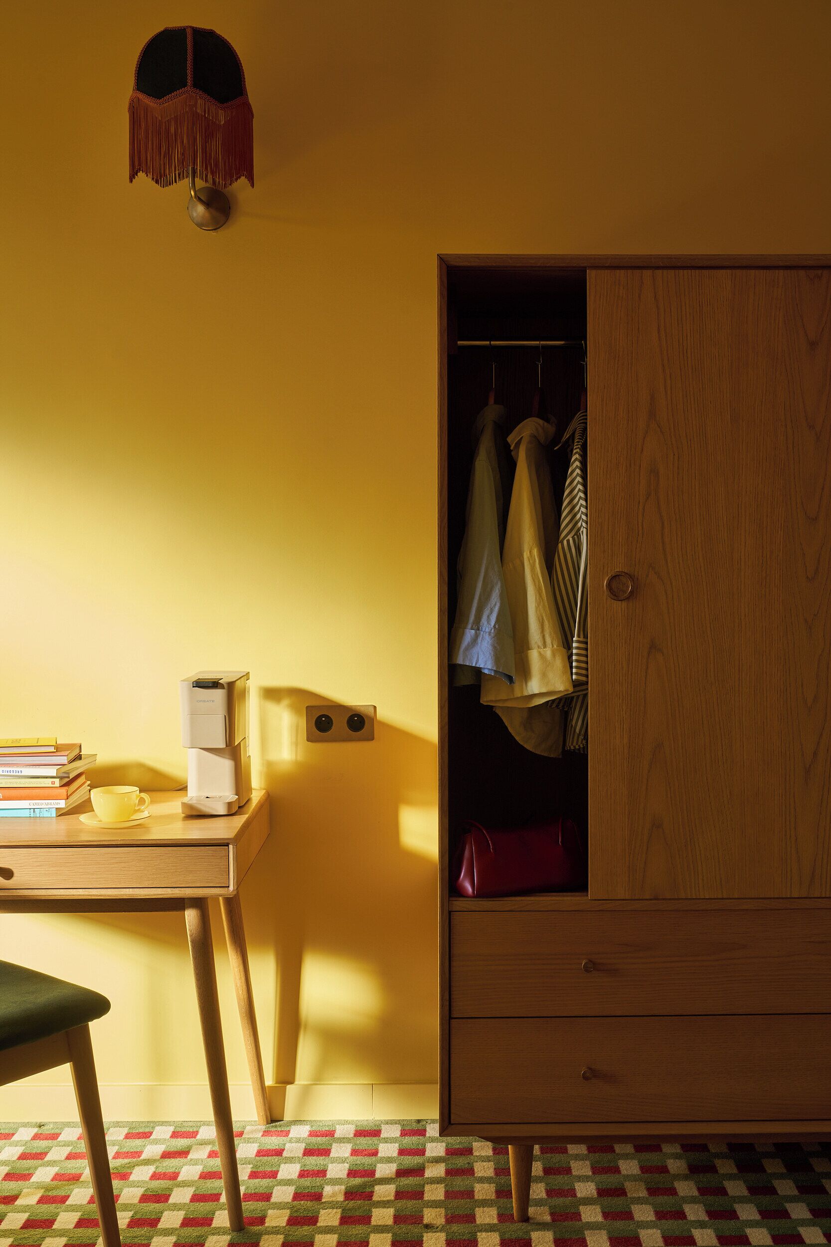 Una habitación con un escritorio de madera, cafetera, una silla, armario y alfombra estampada.