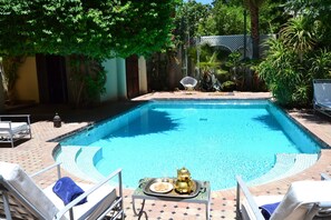 Una piscina con una zona de estar hundida, rodeada de sillas de descanso y una mesa con una bandeja de comida y una tetera.