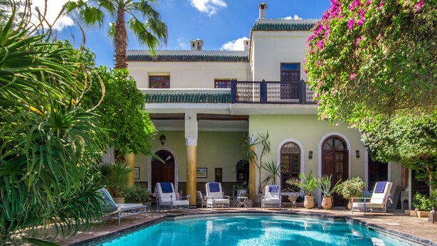 Un área de piscina con sillas de descanso, una piscina cristalina y un edificio con arcos y balcones.