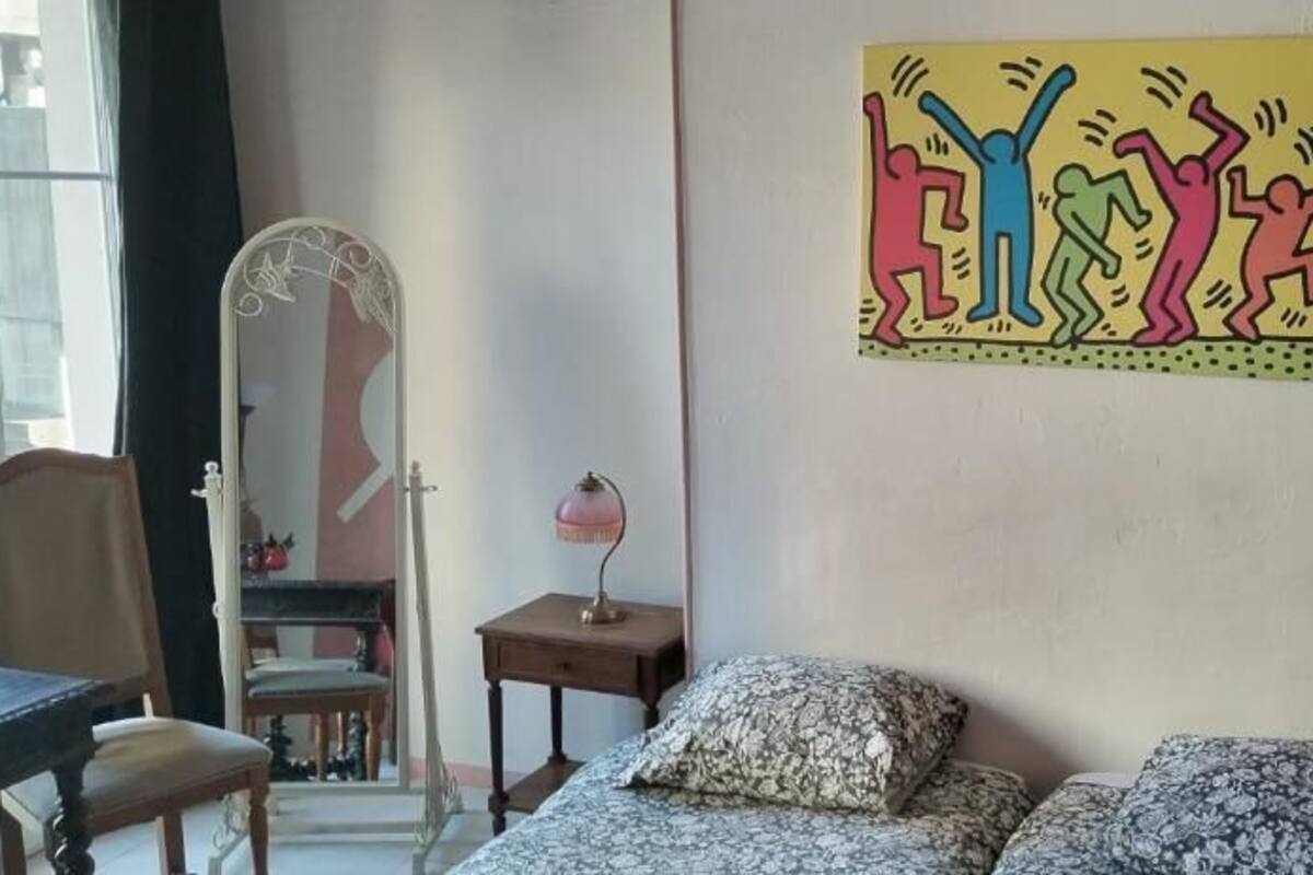 A bedroom with a bed, a chair, a desk, a mirror, and a wall with a colorful artwork.