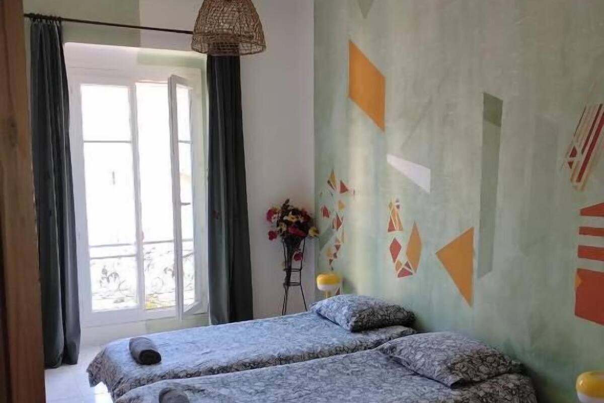 A room with two beds, a hanging light fixture, a small table with a lamp, and a window with curtains.