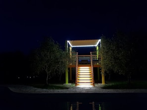 Vista nocturna de un edificio con una escalera que conduce a un área cubierta, rodeado de árboles y una masa de agua.