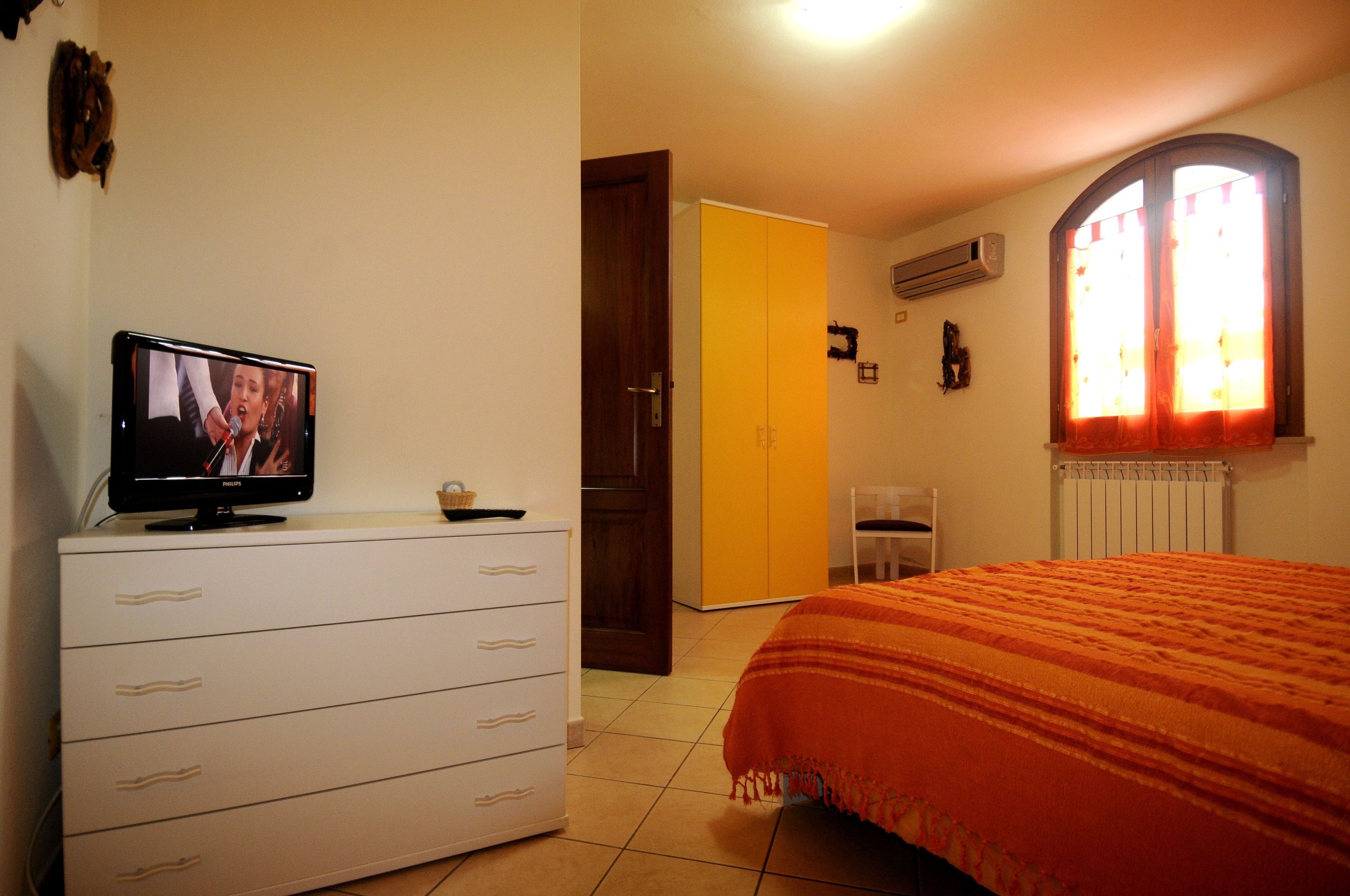 Un dormitorio con cama, televisor sobre una cómoda, ventana con cortinas y una puerta.