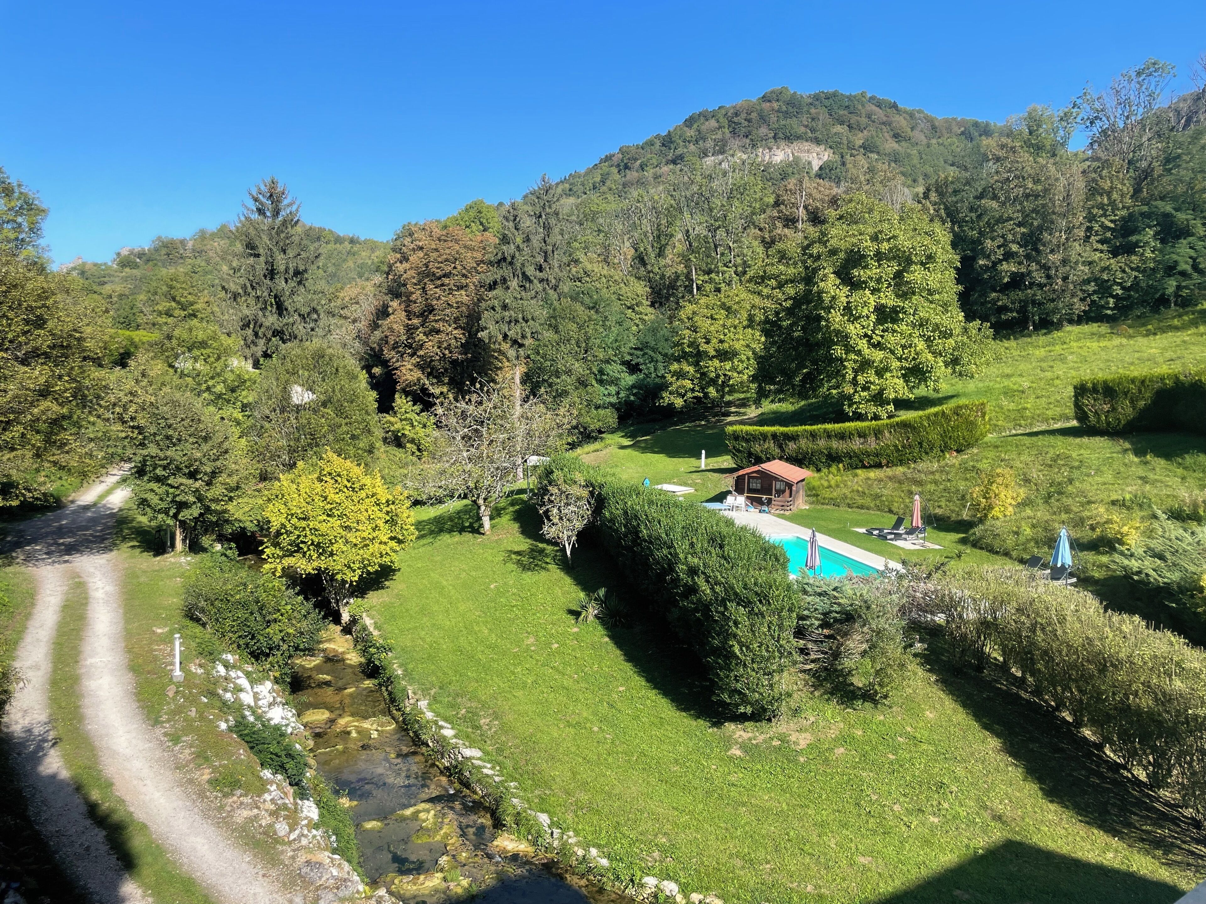 Un paysage verdoyant et luxuriant, avec un chalet en bois, une piscine et un sentier de terre.