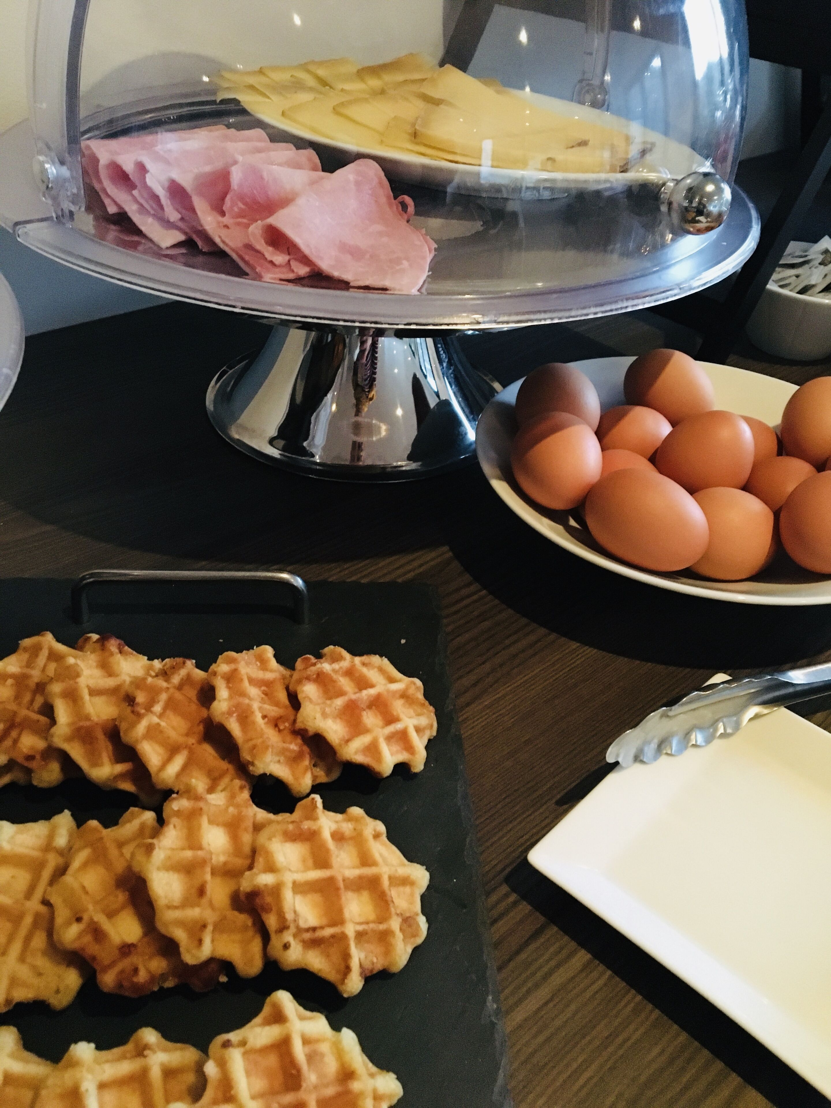 Una mesa de buffet con una variedad de alimentos, entre ellos jamón en rodajas, queso, huevos y gofres.