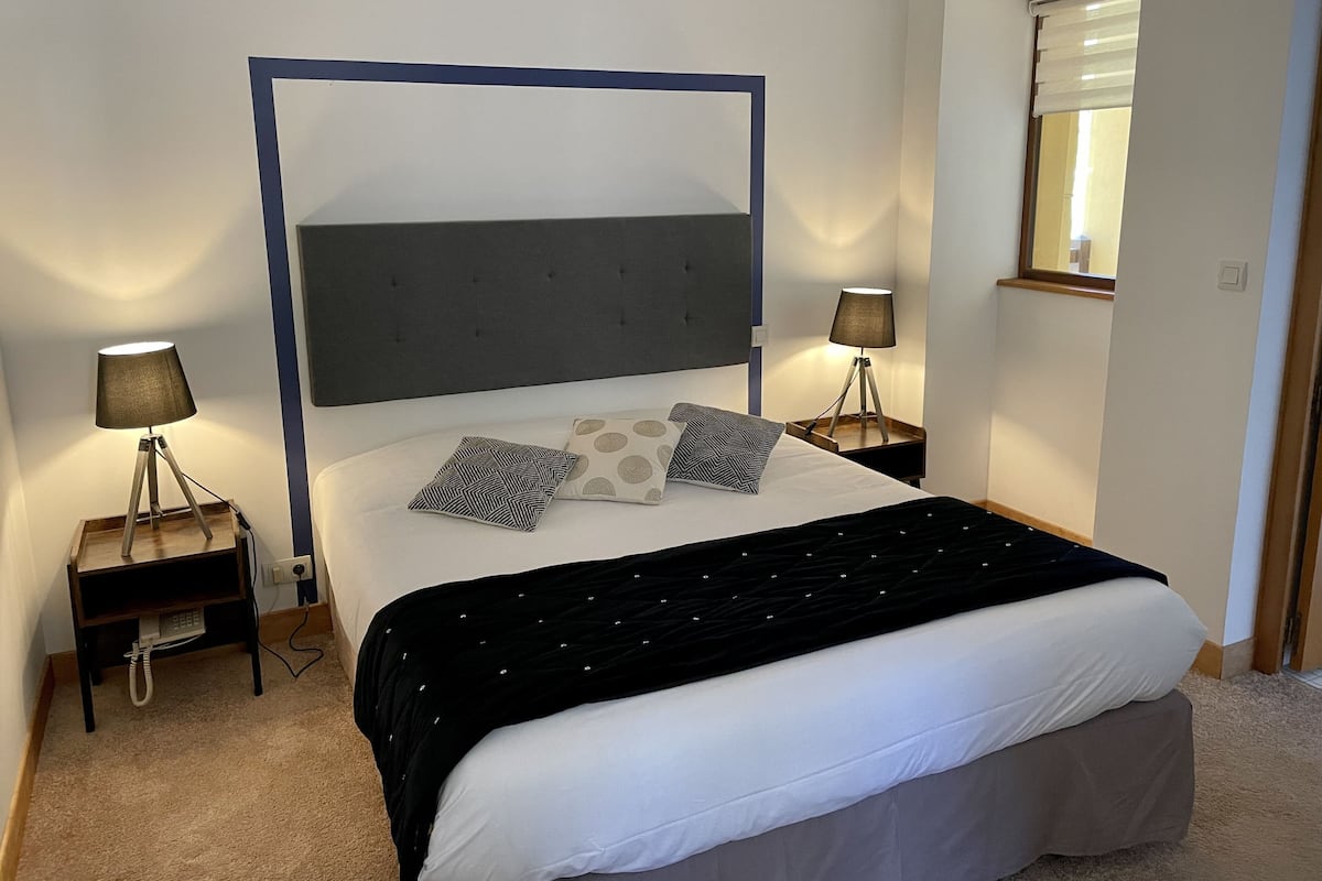 Um quarto com cama, duas mesas de cabeceira, uma lâmpada em cada mesa, janela com persianas e uma porta.