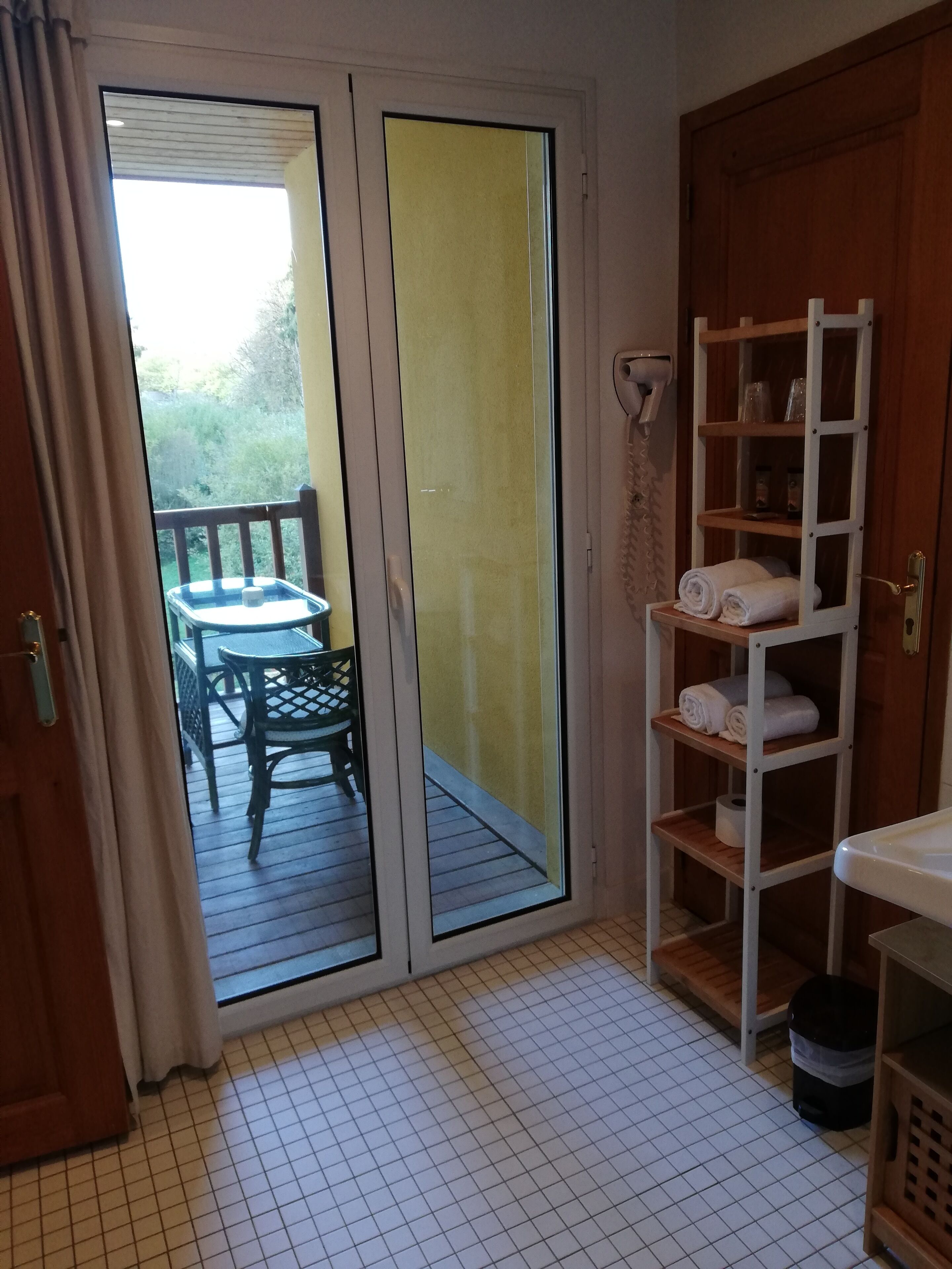 Quarto com porta de vidro deslizante que dá para uma varanda com mesa e cadeiras. Há uma estante de madeira com toalhas e artigos de higiene pessoal.