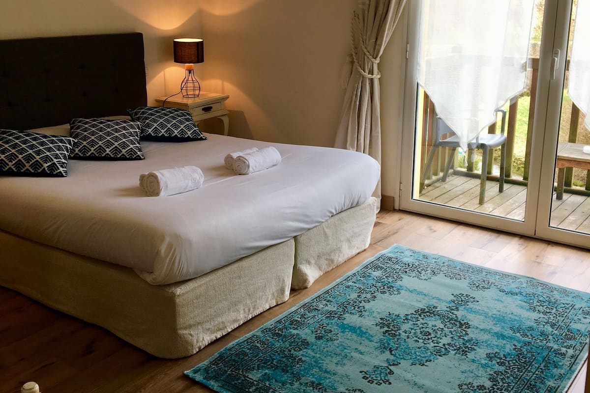 Quarto com uma cama grande, mesa de cabeceira com abajur, quadro na parede e vista para o exterior através de uma porta de vidro deslizante.