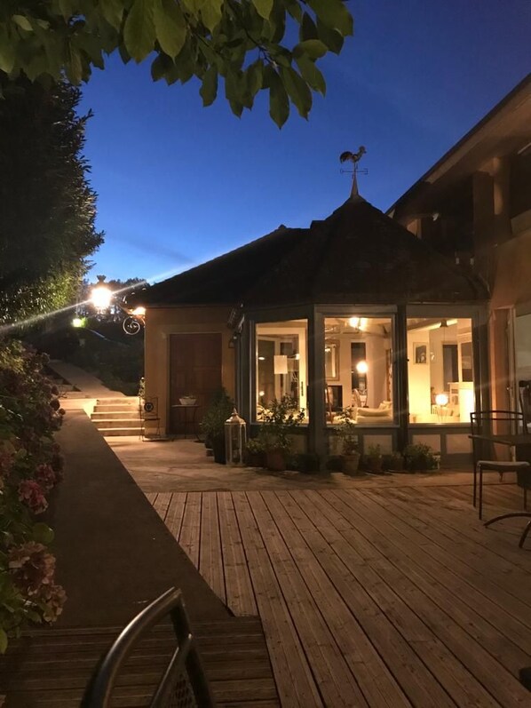 Una terraza de madera que conduce a un edificio con un espacio al aire libre cubierto, una escalera visible y una veleta en el techo.
