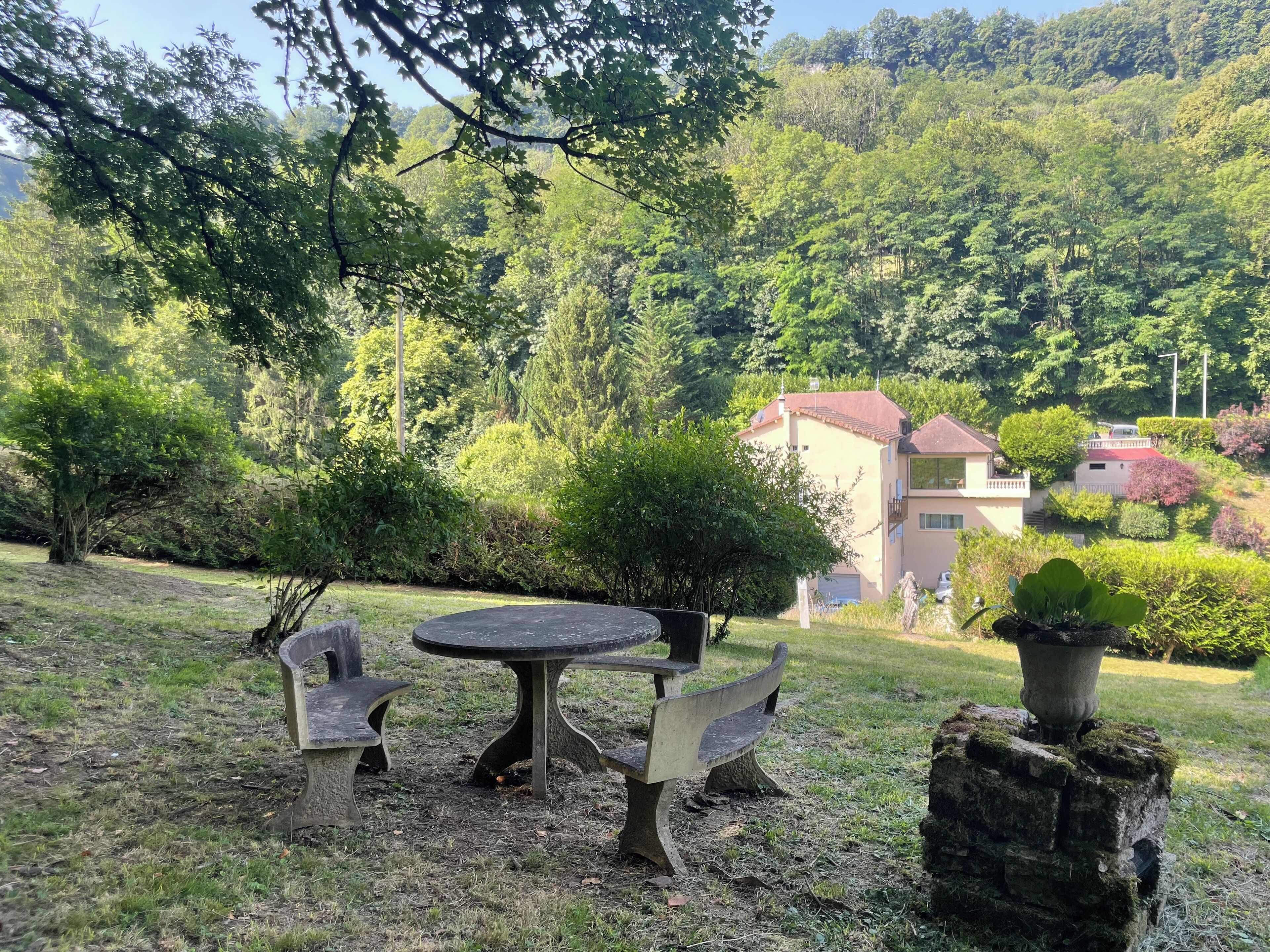 A stone table and benches in a grassy area with a view of a hillside and a building with a red roof.