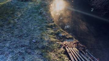 Un cadre naturel en plein air, avec une terrasse en bois, une petite table et des chaises, le tout entouré d’arbres et bordé d’une étendue d’eau qui reflète la lumière du soleil.