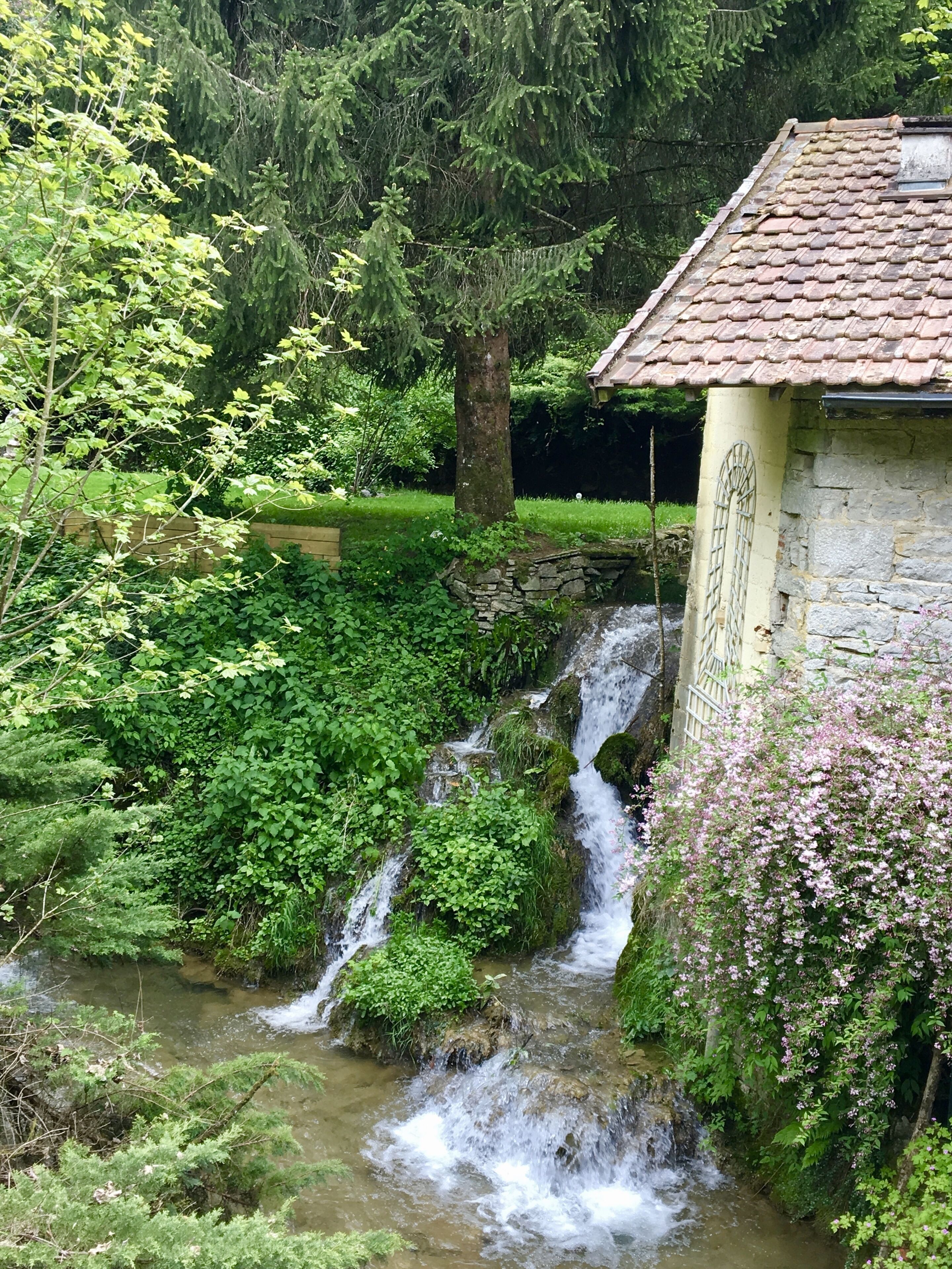 Un edificio de piedra con techo de tejas, una pequeña cascada y abundante vegetación.