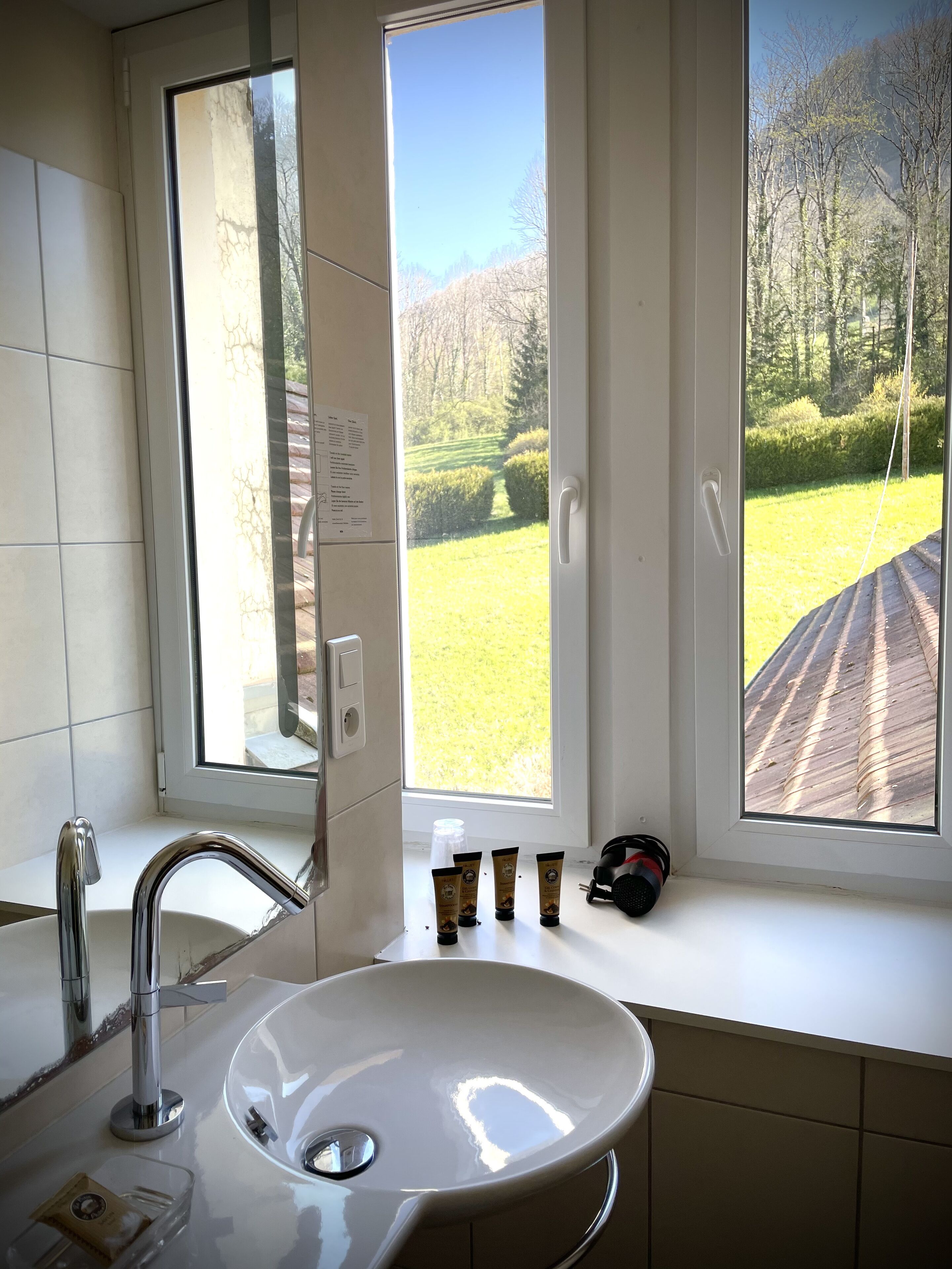 A bathroom with a white sink, a mirror, and a window with a view of a green landscape and trees.