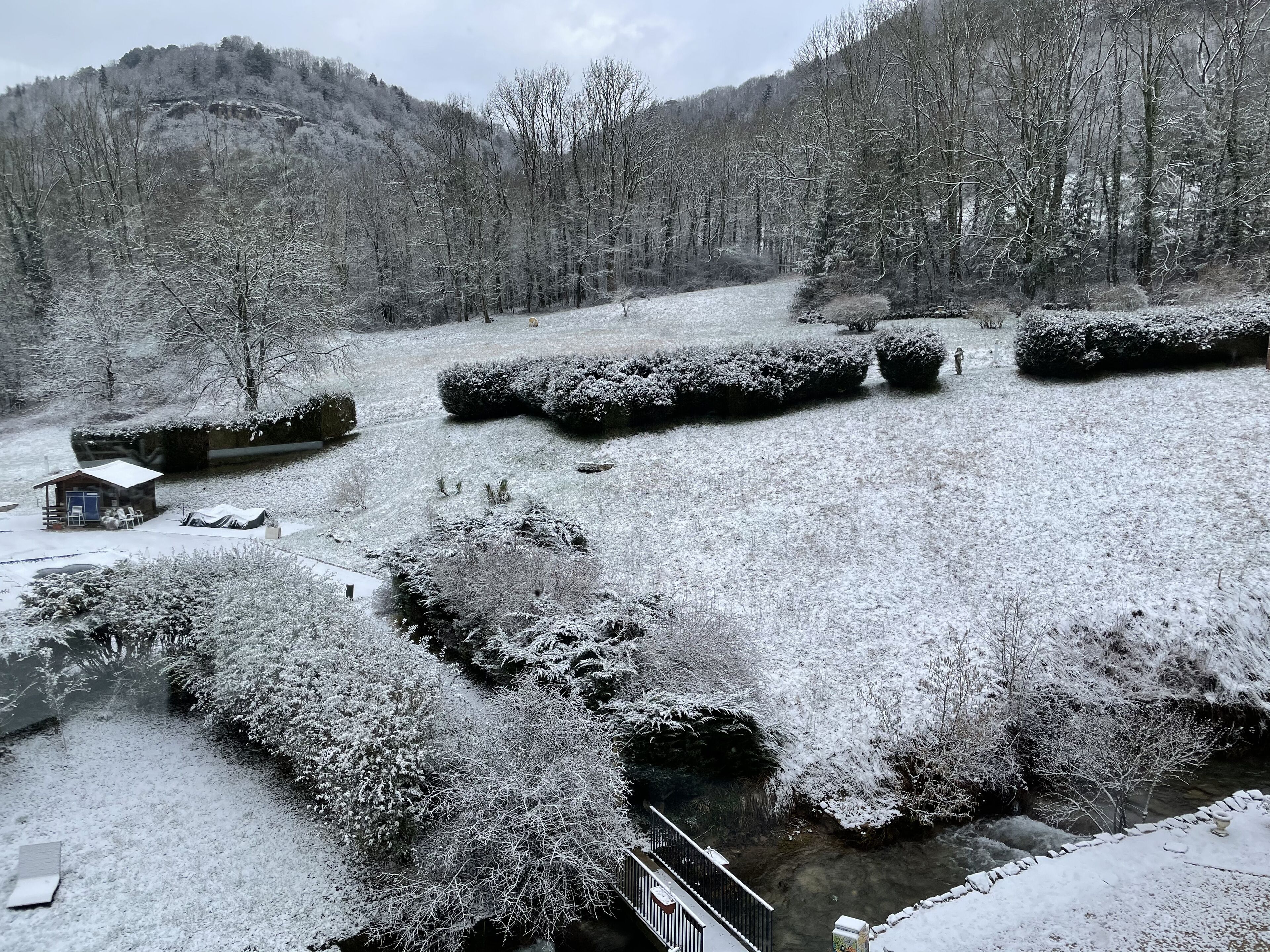 A snow-covered landscape with a cabin, bushes, and a stream.
