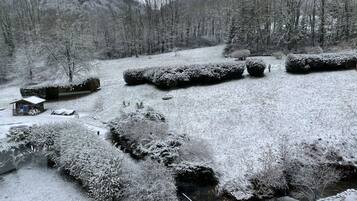 A snow-covered landscape with a cabin, bushes, and a stream.