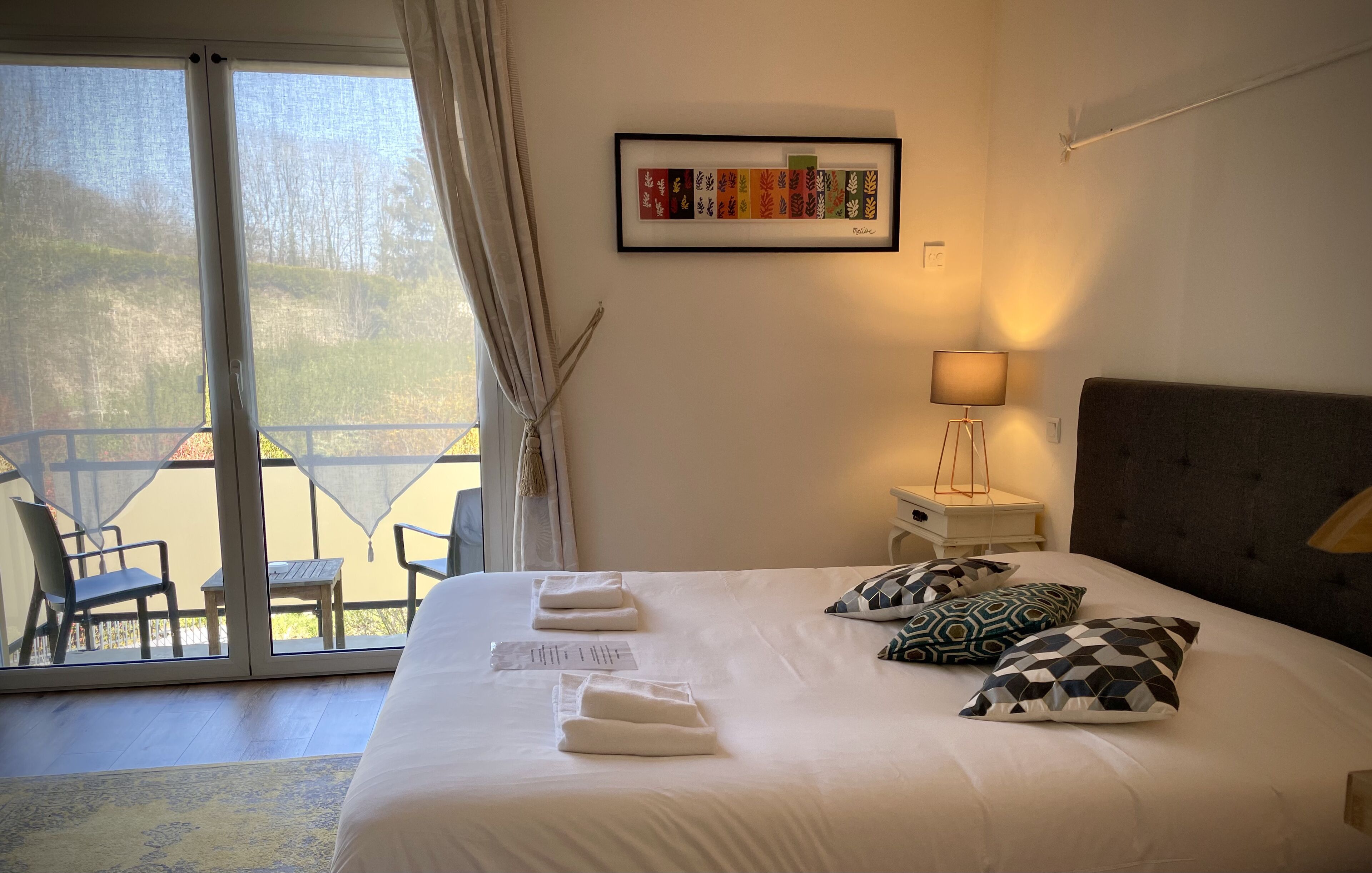 Habitación de hotel con una cama grande, una obra de arte enmarcada en la pared, una mesita de noche con lámpara y un balcón con sillas.
