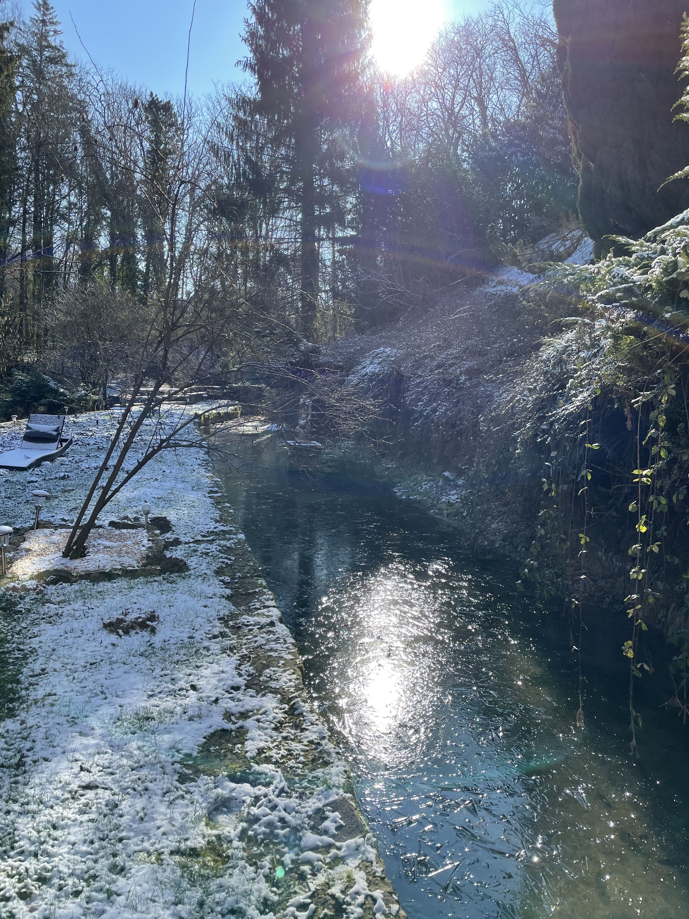Escena apacible a orillas del río, con suelo y árboles nevados, reflejando la luz del sol en el agua.