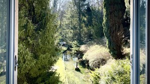 A view through a window of a forested area with a stream, trees, and a statue. - Hôtel de la Vallée Heureuse (Poligny)