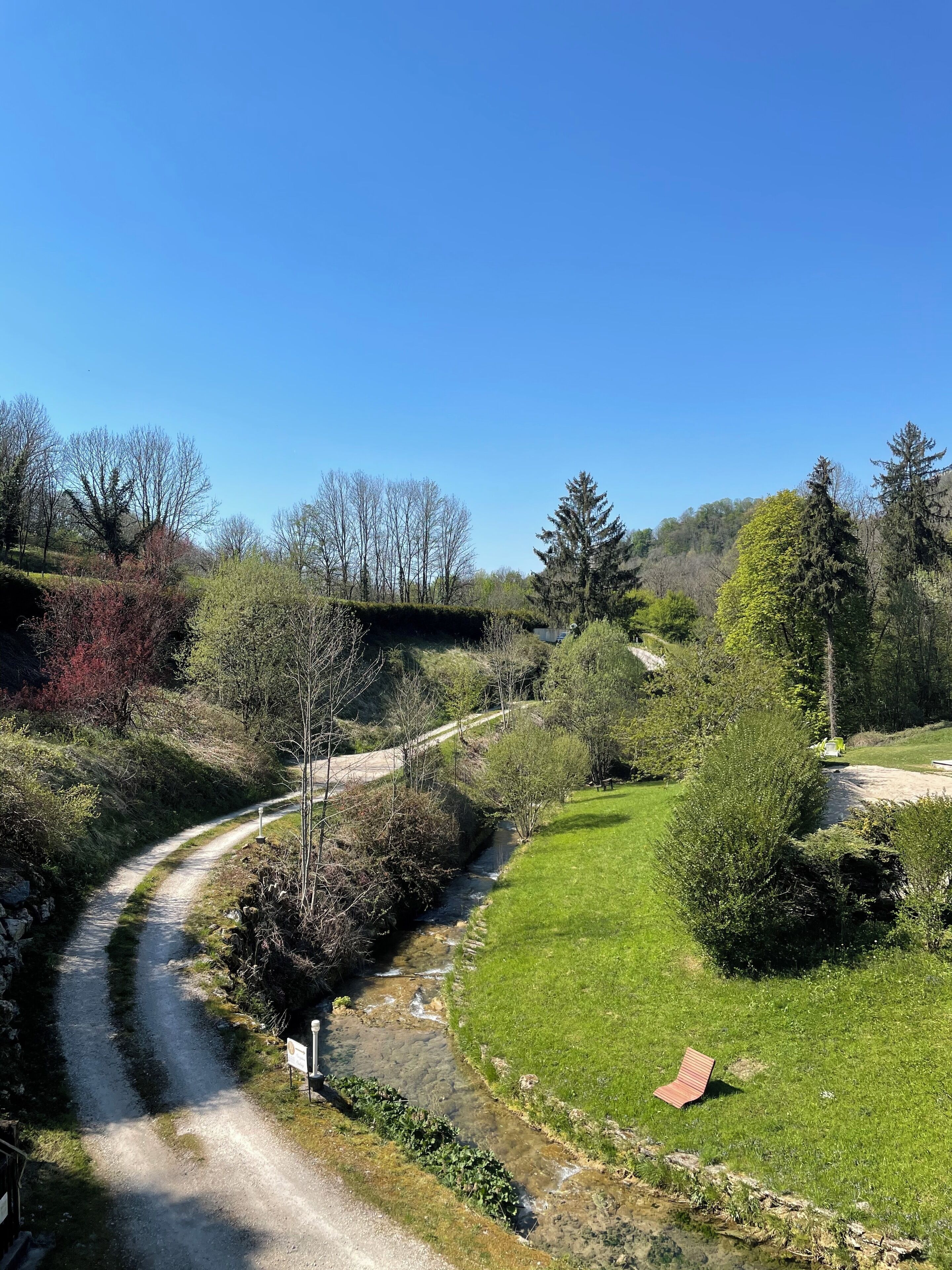 A rural landscape with a winding dirt path, a small stream, and a bench.