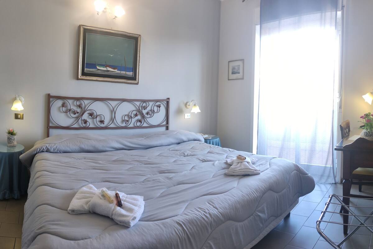 A neatly made bed with a white duvet, a small bedside table with a lamp, a framed picture on the wall, and a window with curtains.