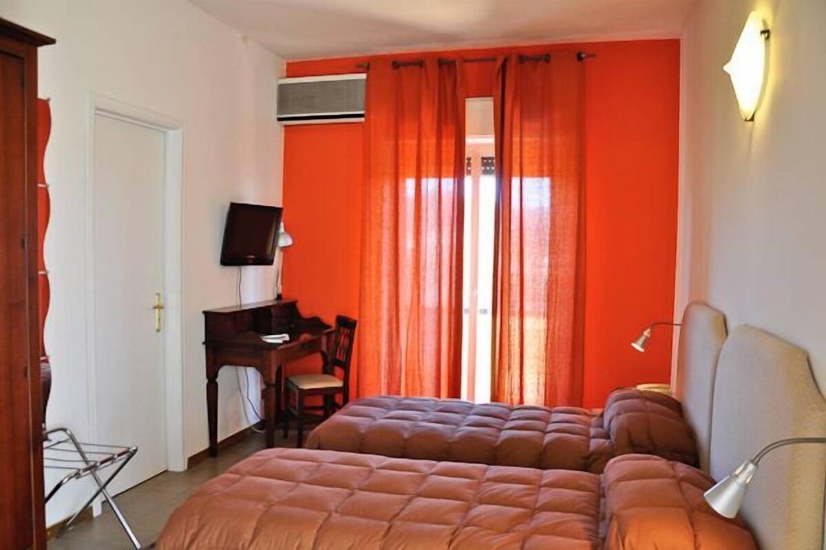 A bedroom with two beds, a desk with a chair, a television, and a window with curtains.