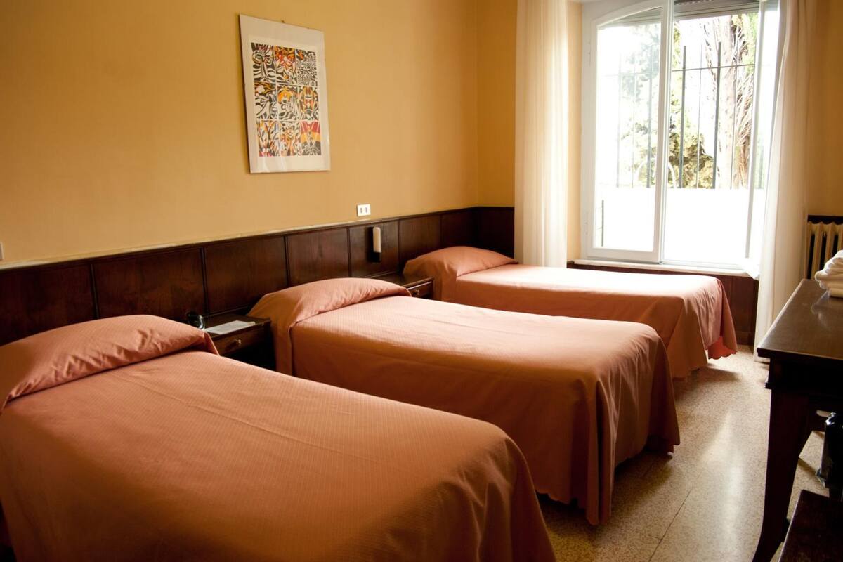 A hotel room with two beds, a wooden headboard, a window with a view of greenery, and a table with a lamp.