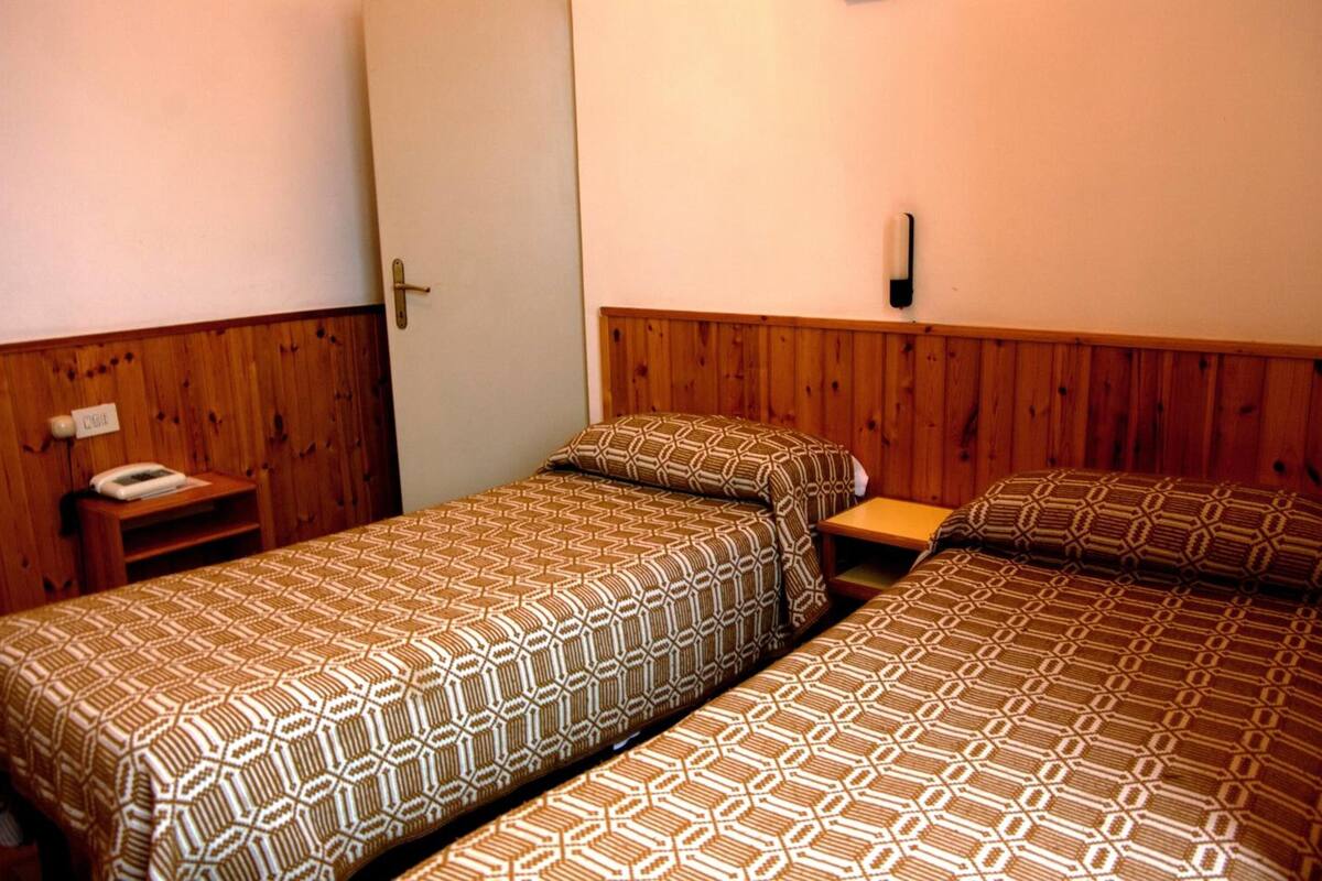 A room with two beds, wooden paneling on the walls, a bedside table with a phone, and a wall-mounted light.