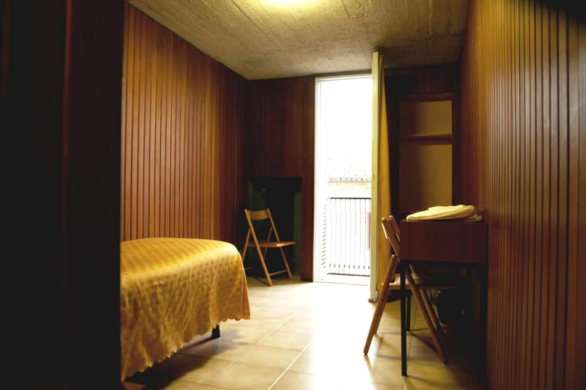 A small, cozy room with wooden walls and a single bed. There is a chair and a small table with a chair. A door leads to an outdoor area.