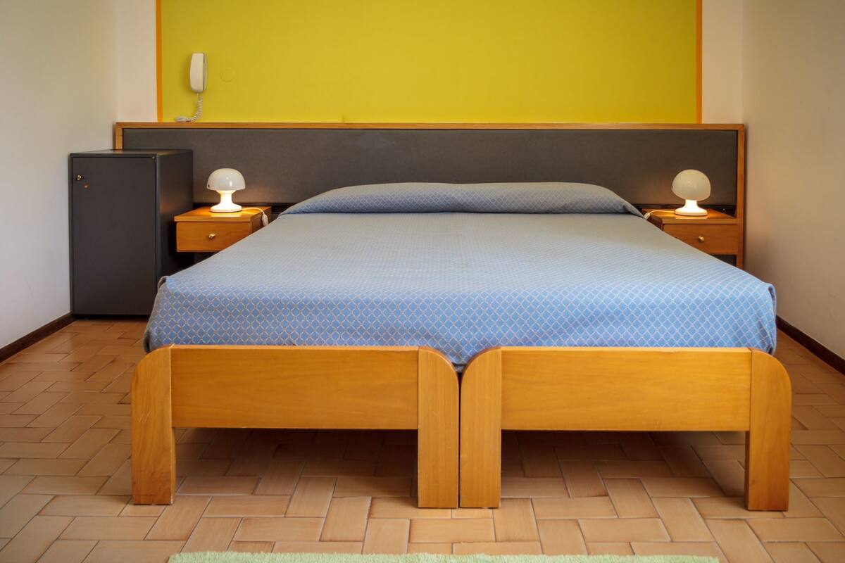 A single bed with a wooden frame and a blue mattress, flanked by two bedside tables with lamps, in a room with a herringbone floor pattern.