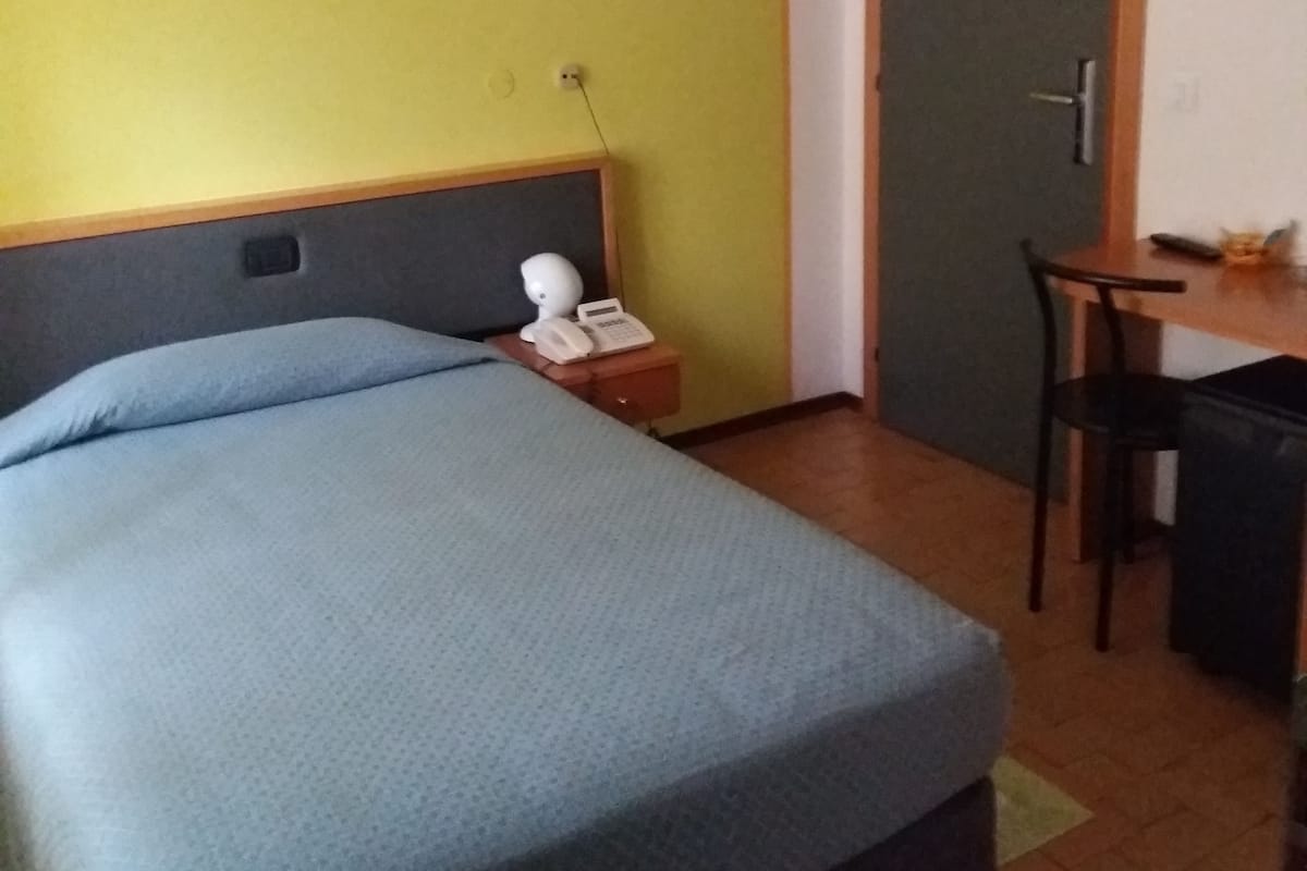 A hotel room with a bed, a desk, a chair, a telephone, and a door.