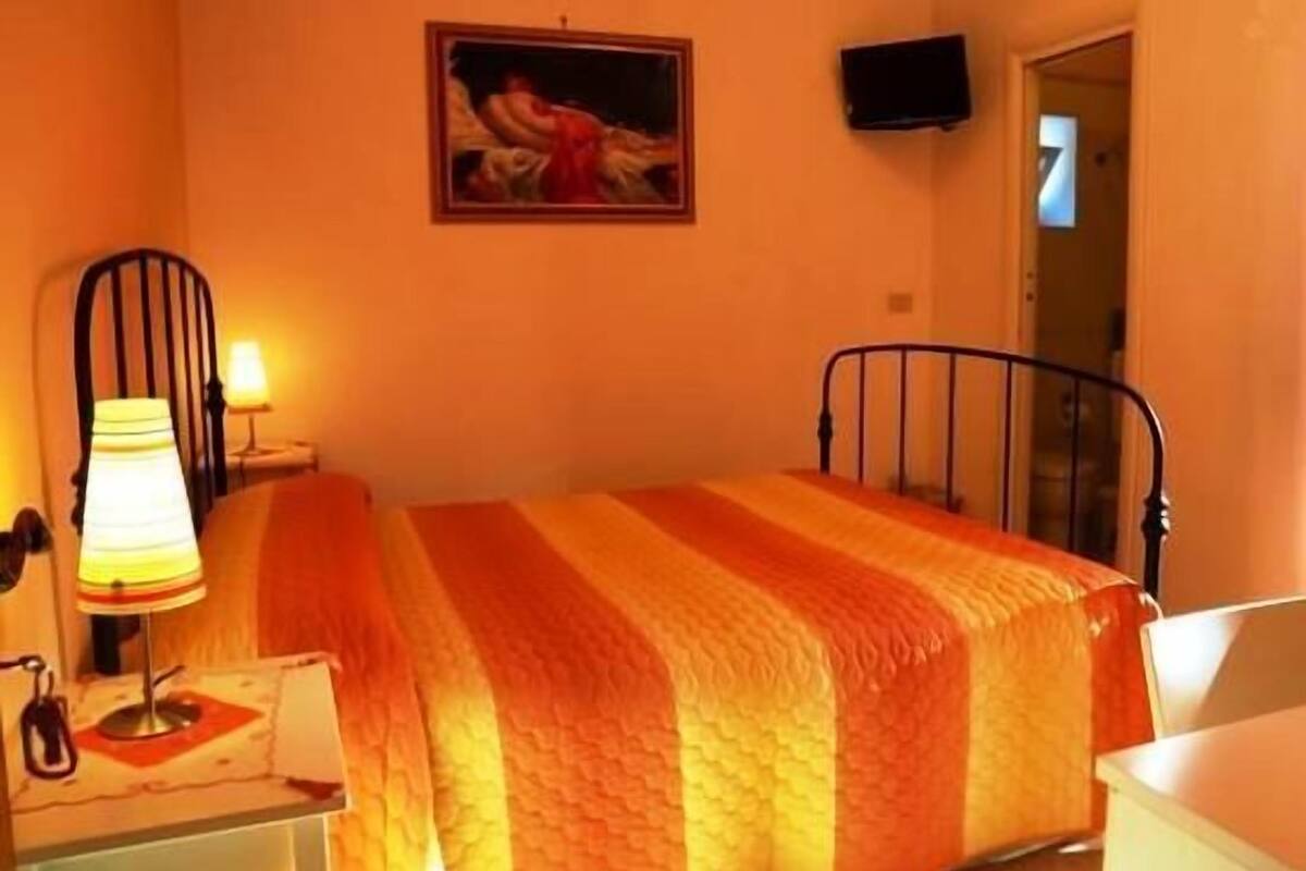 A bedroom with a bed, a lamp, a painting, and a television on the wall.