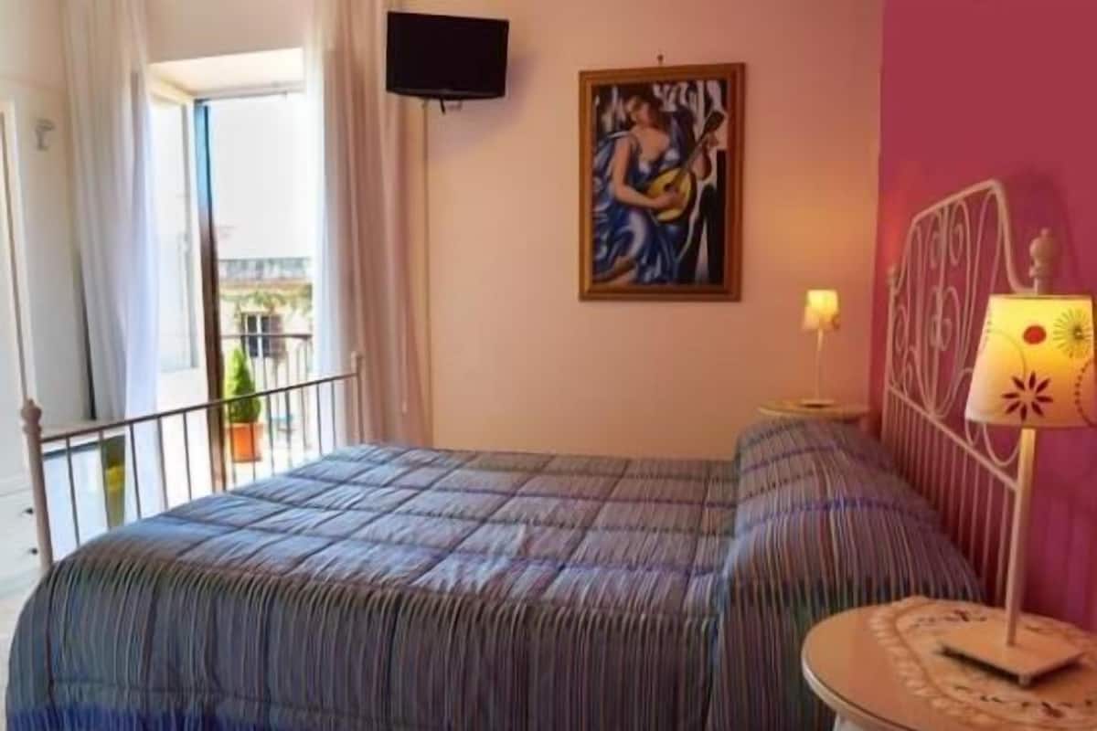 A room with a bed, a painting on the wall, a TV mounted above the bed, and a window with curtains.